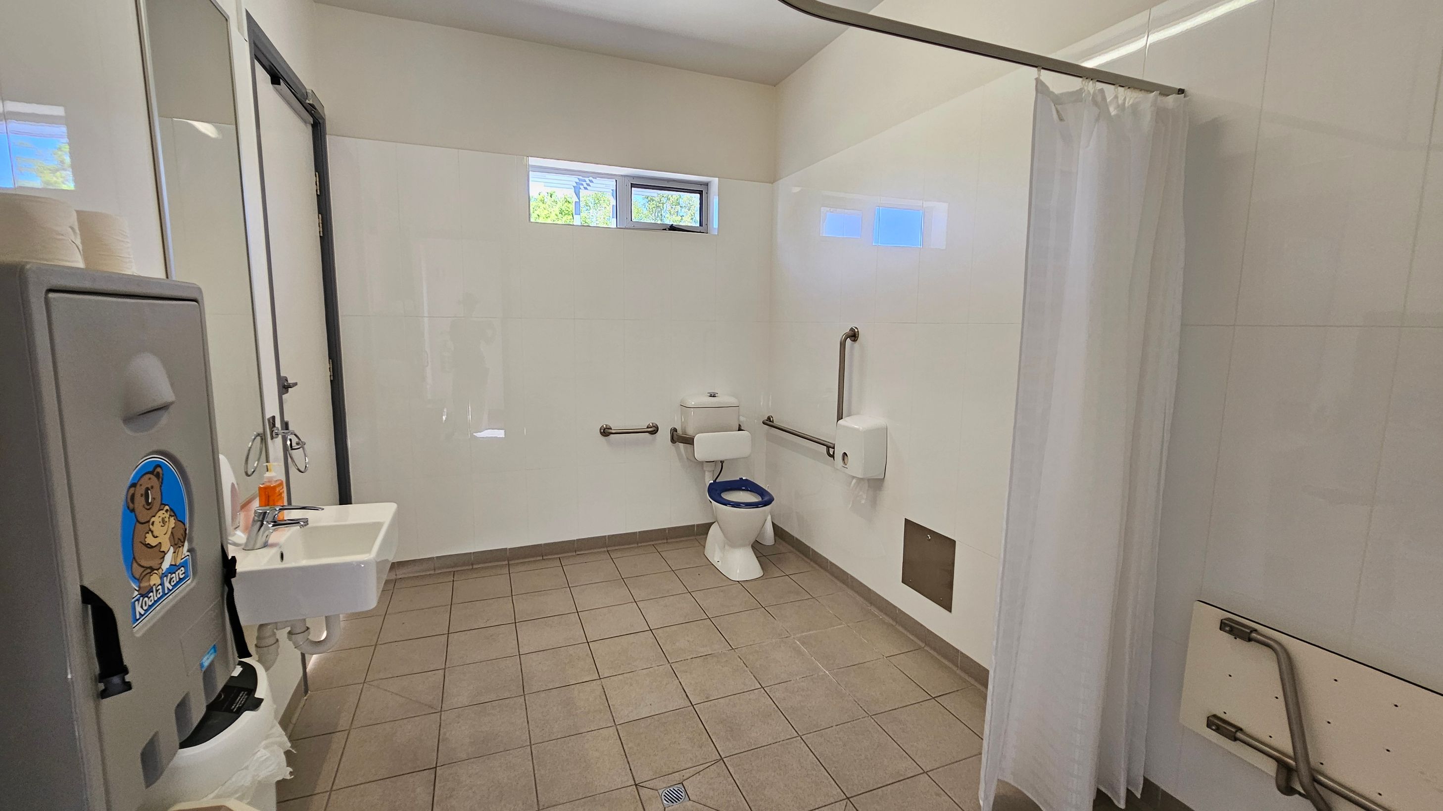 A public restroom with a tiled floor and light-coloured walls. On the left, there's a baby changing station with a "Koala Kare" logo, a sink with a soap dispenser, and a mirror. In the centre, a toilet with a blue seat is equipped with grab bars on both sides. On the right, a white shower curtain is partially visible. High windows near the ceiling allow natural light in.