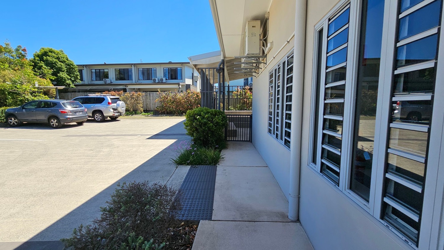 A paved area with a building on the right featuring large windows with horizontal slats. In the foreground, there are small bushes and plants along the path. On the left, a parking area with two cars, a grey and a white one, is visible. In the background, a two-storey building with air conditioning units on the upper level and a wooden fence surrounded by greenery. The sky is clear and blue.