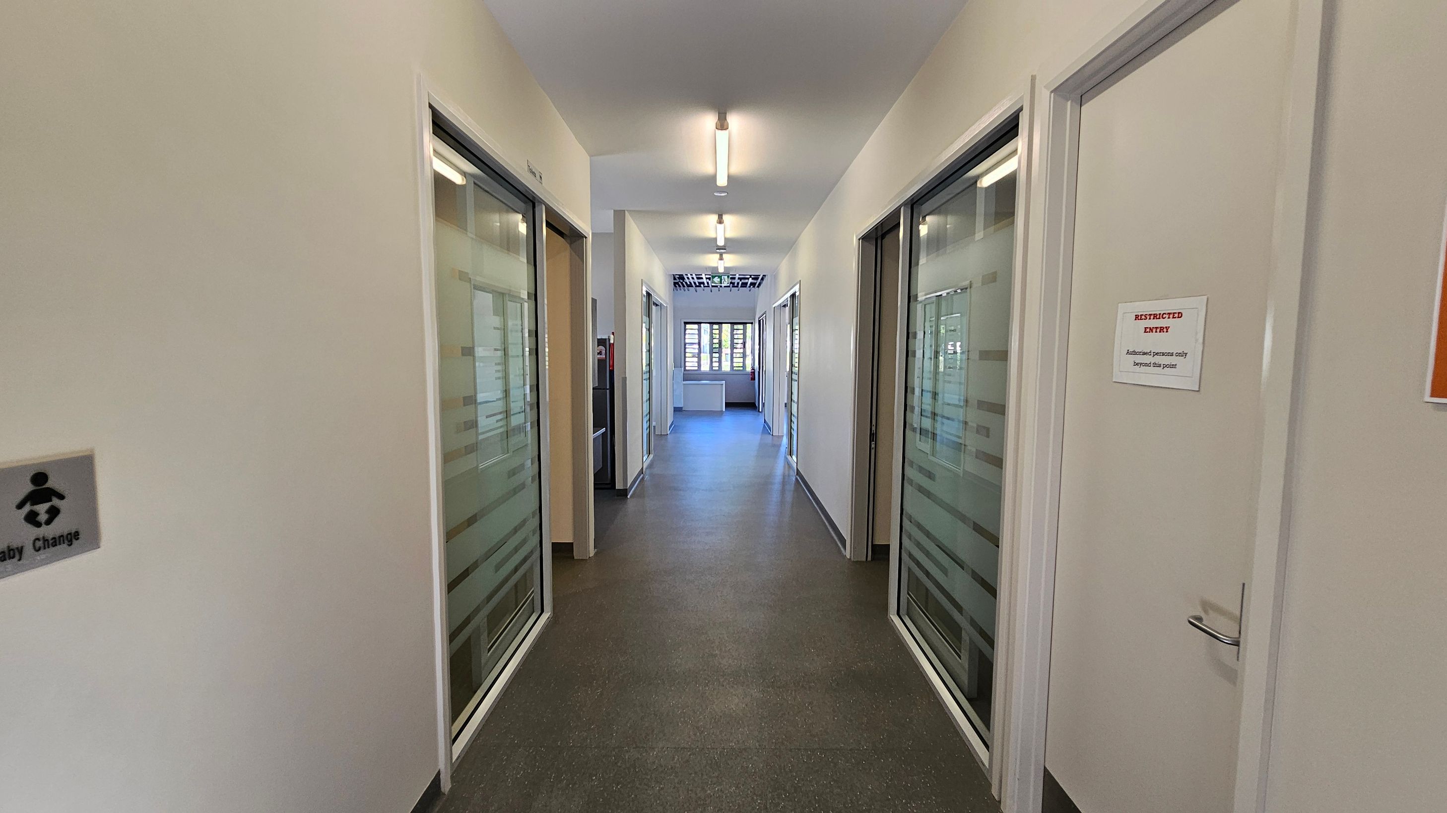 A corridor with a grey speckled floor and white walls. On the left, there's a sign for a baby changing area. On the right, a door with a "Restricted Entry" sign. Both sides have glass-panelled doors with frosted stripes. The ceiling has fluorescent lights, and at the end of the corridor, there's a window with horizontal blinds letting in natural light.