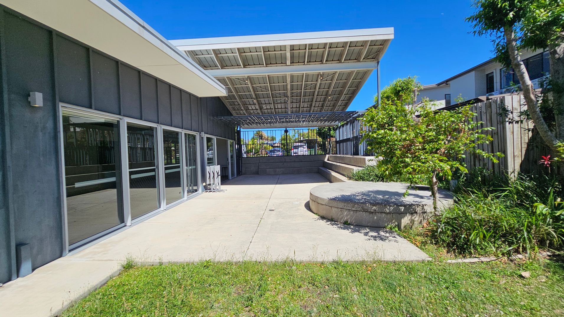 A modern outdoor area with a concrete patio and a grassy foreground. On the left, there is a building with large glass doors and a dark exterior. In the centre, a large metal canopy provides shade. To the right, there are raised concrete seating areas surrounded by greenery, including small trees and plants. A wooden fence and neighbouring buildings are visible in the background.