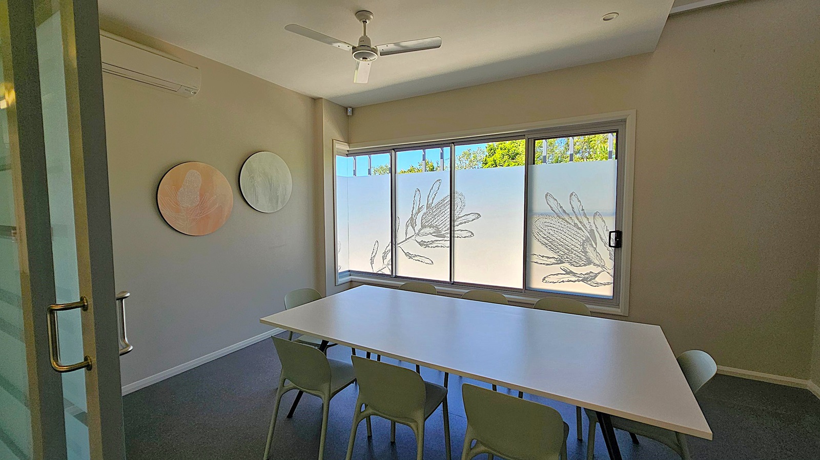 A small meeting room with a white rectangular table surrounded by six chairs. On the right wall, a flat-screen TV is mounted. The left side shows a glass door with a metal handle. The back wall features large windows with frosted glass and decorative plant designs, allowing some natural light. The ceiling has a white fan, and the floor is carpeted in a dark colour.