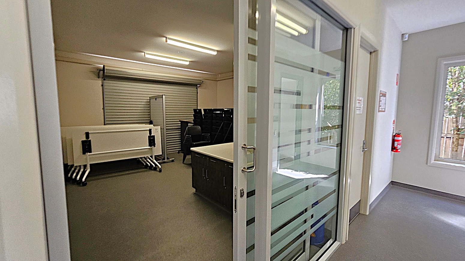 A room with a glass sliding door on the right, featuring horizontal frosted stripes. Inside, there are stacked tables and chairs against a wall with a closed roller shutter. The floor is grey, and the ceiling has fluorescent lights. In the hallway, a fire extinguisher is mounted on the wall near a window with a view of trees and a wooden fence outside.