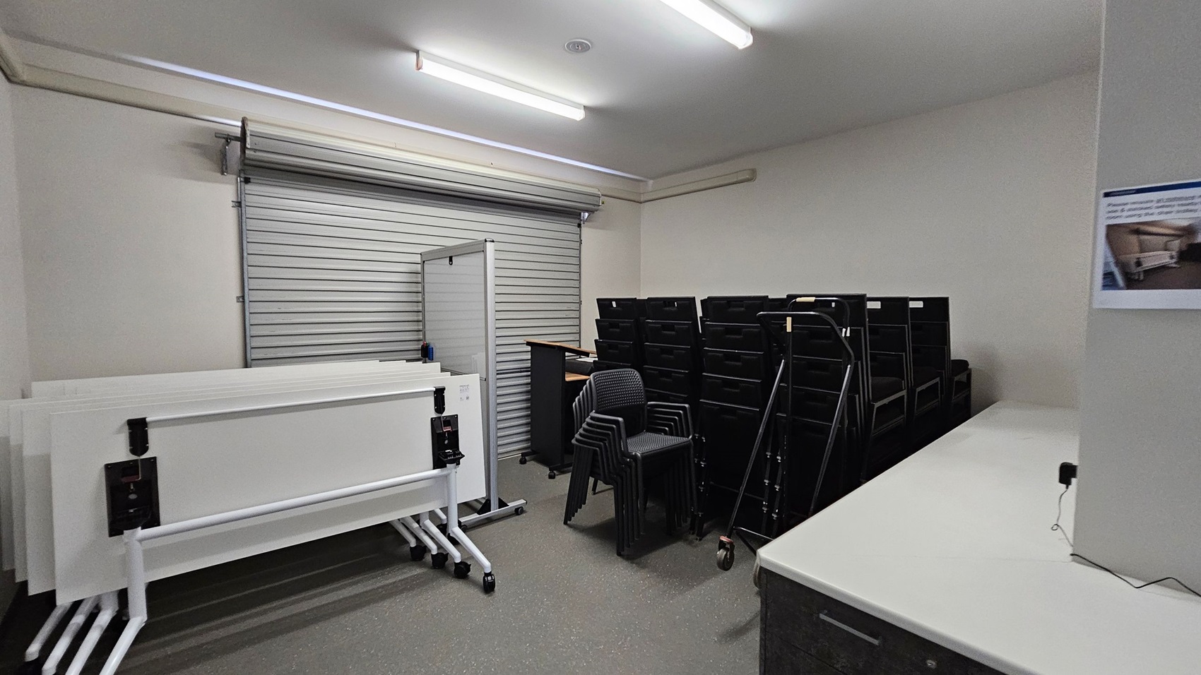 A storage room with a grey roller shutter door on the left. In front of it, several white folding tables are stacked on wheeled frames. In the centre, there are stacked black chairs and a trolley. On the right, a row of black storage units is visible. A white countertop runs along the right wall, with a poster above it. The floor is grey and the ceiling has fluorescent lights.