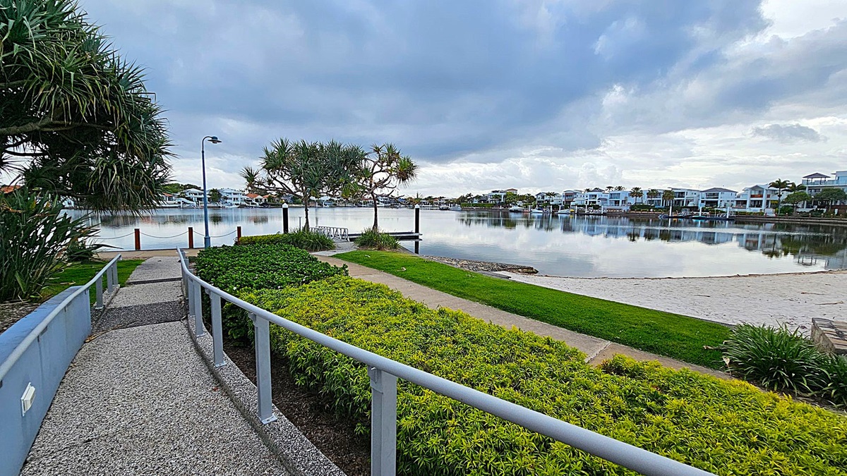 A paved path leads through a landscaped area with neatly trimmed bushes and palm trees. On the left, there is a modern building with a flat roof and a small set of stairs. On the right, a ramp with a metal railing ascends towards another building partially obscured by trees. The foreground features a mix of grass and mulch, while the background includes more greenery and architectural elements. A paved path leads through a landscaped area with neatly trimmed bushes and palm trees. On the left, there is a modern building with a flat roof and a small set of stairs. On the right, a ramp with a metal railing ascends towards another building partially obscured by trees. The foreground features a mix of grass and mulch, while the background includes more greenery and architectural elements.