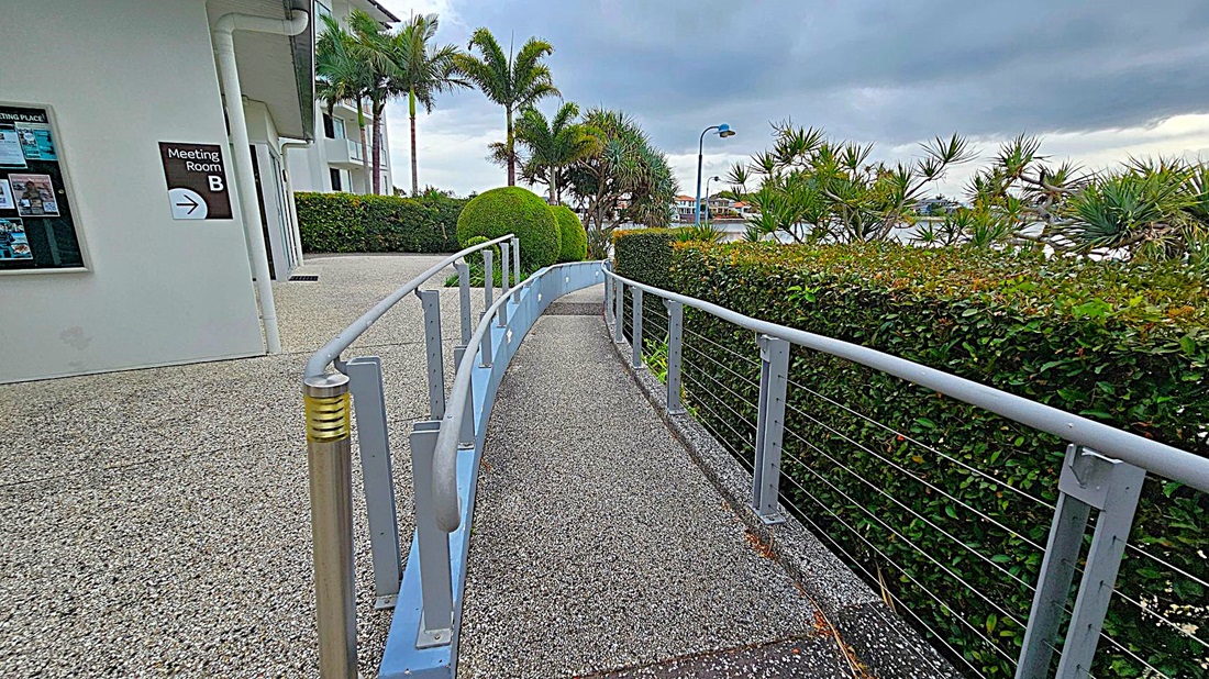 A pathway with a grey, speckled surface curves gently to the right, bordered by a metal railing. On the left, a building with a sign for "Meeting Room B" and a notice board is visible. To the right, a neatly trimmed hedge runs alongside the path, with palm trees and other greenery in the background. A streetlamp and a glimpse of water and buildings can be seen in the distance. A pathway with a grey, speckled surface curves gently to the right, bordered by a metal railing. On the left, a building with a sign for "Meeting Room B" and a notice board is visible. To the right, a neatly trimmed hedge runs alongside the path, with palm trees and other greenery in the background. A streetlamp and a glimpse of water and buildings can be seen in the distance.