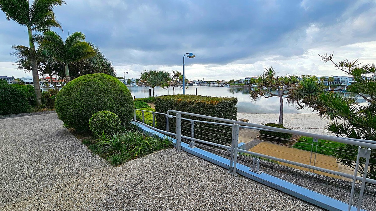 A waterfront scene with a pebble pathway in the foreground, bordered by neatly trimmed bushes and a round, manicured shrub on the left. Tall palm trees rise behind the shrubbery. A metal railing runs along the path, overlooking a calm body of water. Across the water, modern houses are visible. The sky is overcast, casting a grey tone over the landscape. A waterfront scene with a pebble pathway in the foreground, bordered by neatly trimmed bushes and a round, manicured shrub on the left. Tall palm trees rise behind the shrubbery. A metal railing runs along the path, overlooking a calm body of water. Across the water, modern houses are visible. The sky is overcast, casting a grey tone over the landscape.