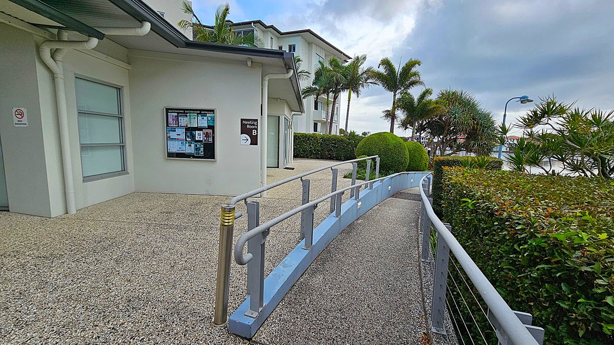 A building with a grey exterior and a window on the left. A notice board and a sign for "Meeting Room B" are mounted on the wall. A ramp with metal railings curves from the foreground to the right, bordered by a hedge. Palm trees and manicured bushes are visible in the background, along with a streetlamp. The ground is covered with a speckled surface. A building with a grey exterior and a window on the left. A notice board and a sign for "Meeting Room B" are mounted on the wall. A ramp with metal railings curves from the foreground to the right, bordered by a hedge. Palm trees and manicured bushes are visible in the background, along with a streetlamp. The ground is covered with a speckled surface.