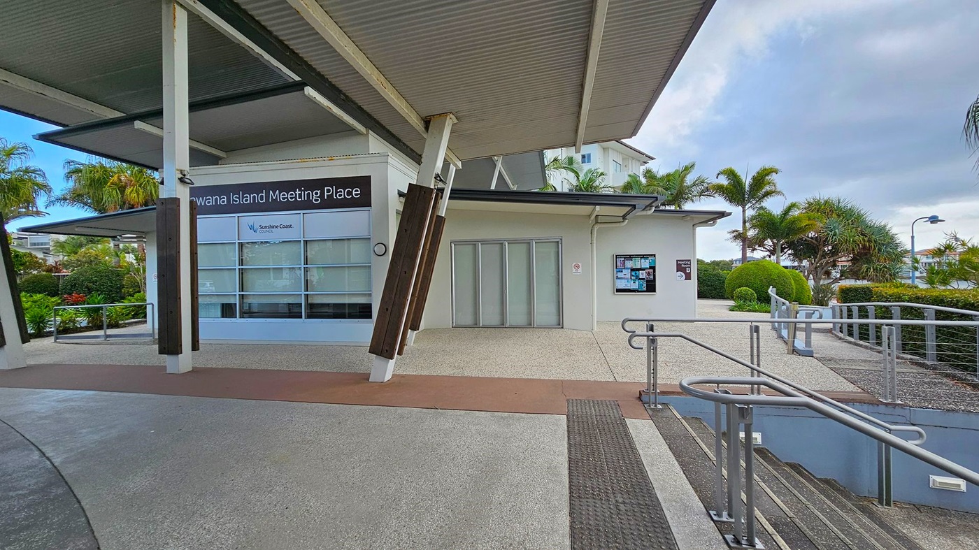 A modern building with a sign reading "Kawana Island Meeting Place" on the left. The building has large windows and a corrugated metal roof. In the centre, there are closed sliding doors and a notice board on the wall. On the right, a pathway with metal railings leads to a staircase. The area is surrounded by well-maintained greenery, including palm trees and bushes. The ground is paved with textured concrete. A modern building with a sign reading "Kawana Island Meeting Place" on the left. The building has large windows and a corrugated metal roof. In the centre, there are closed sliding doors and a notice board on the wall. On the right, a pathway with metal railings leads to a staircase. The area is surrounded by well-maintained greenery, including palm trees and bushes. The ground is paved with textured concrete.