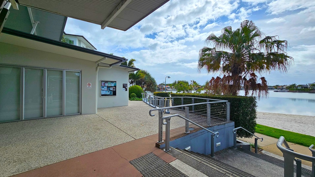 A building with large glass doors on the left, featuring a notice board on the wall. A pathway leads to a railing and steps descending to a lower level. On the right, a palm tree stands near a body of water with a sandy shore. The background includes more trees and distant buildings across the water. The floor is a mix of concrete and textured surfaces, and the ceiling is metal with visible beams. A building with large glass doors on the left, featuring a notice board on the wall. A pathway leads to a railing and steps descending to a lower level. On the right, a palm tree stands near a body of water with a sandy shore. The background includes more trees and distant buildings across the water. The floor is a mix of concrete and textured surfaces, and the ceiling is metal with visible beams.
