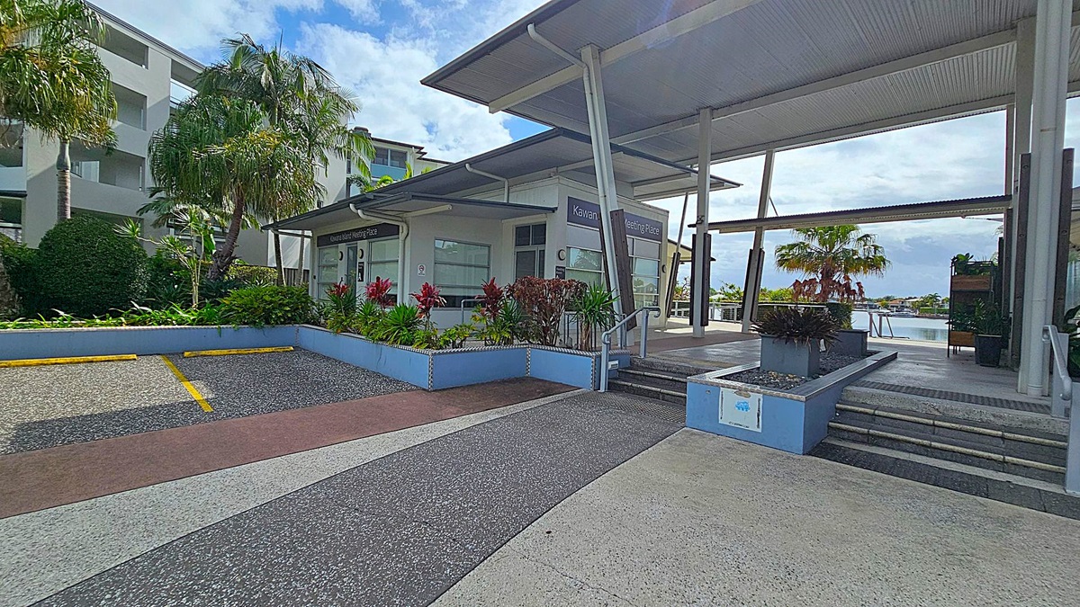 A modern building with a slanted roof and large windows is on the right, labelled "Kawana Island Meeting Place." Steps lead up to the entrance, flanked by planters with red and green foliage. On the left, there are palm trees and a multi-storey building with balconies. The foreground has a textured pavement with yellow parking lines. In the background, there's a glimpse of water and more greenery. A modern building with a slanted roof and large windows is on the right, labelled "Kawana Island Meeting Place." Steps lead up to the entrance, flanked by planters with red and green foliage. On the left, there are palm trees and a multi-storey building with balconies. The foreground has a textured pavement with yellow parking lines. In the background, there's a glimpse of water and more greenery.
