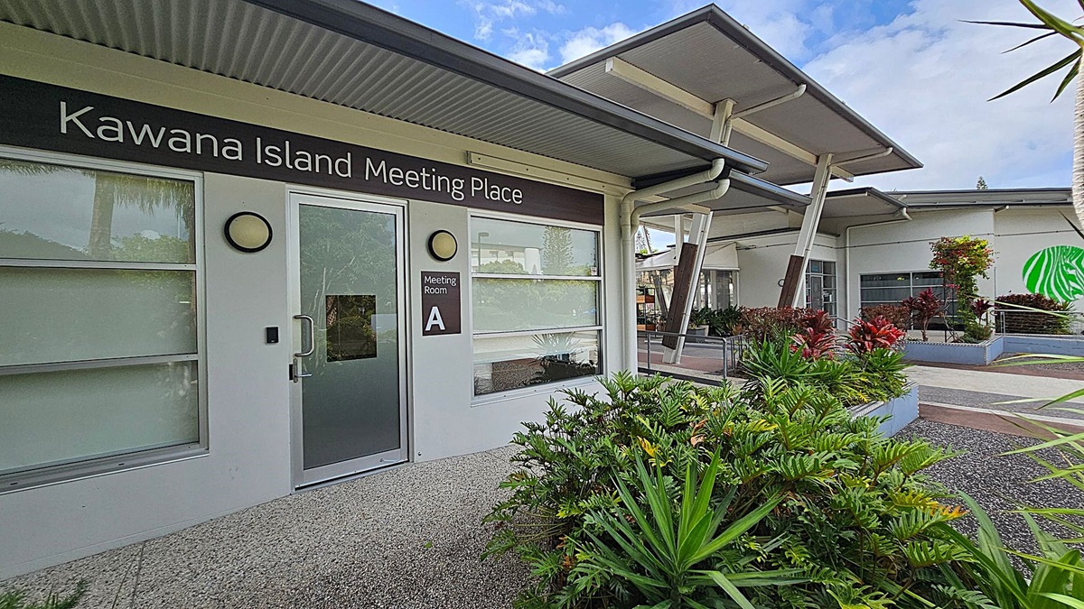 A modern building with a sign reading "Kawana Island Meeting Place" above a door labelled "Meeting Room A". The exterior features large windows and a grey, corrugated roof. In the foreground, there are lush green plants and a textured pathway. To the right, more plants are in raised beds, and a green abstract design is visible on a distant wall. The structure has a contemporary design with angled supports and overhangs. A modern building with a sign reading "Kawana Island Meeting Place" above a door labelled "Meeting Room A". The exterior features large windows and a grey, corrugated roof. In the foreground, there are lush green plants and a textured pathway. To the right, more plants are in raised beds, and a green abstract design is visible on a distant wall. The structure has a contemporary design with angled supports and overhangs.