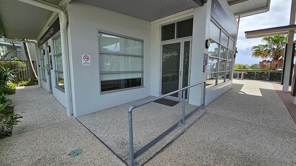 A building corner with large windows and a glass door. A "No Smoking" sign is on the wall. A metal handrail runs along the edge of the concrete path. To the left, there are plants and a fence. On the right, a pathway leads to a railing with palm trees and greenery in the background. The ceiling is corrugated metal. A building corner with large windows and a glass door. A "No Smoking" sign is on the wall. A metal handrail runs along the edge of the concrete path. To the left, there are plants and a fence. On the right, a pathway leads to a railing with palm trees and greenery in the background. The ceiling is corrugated metal.