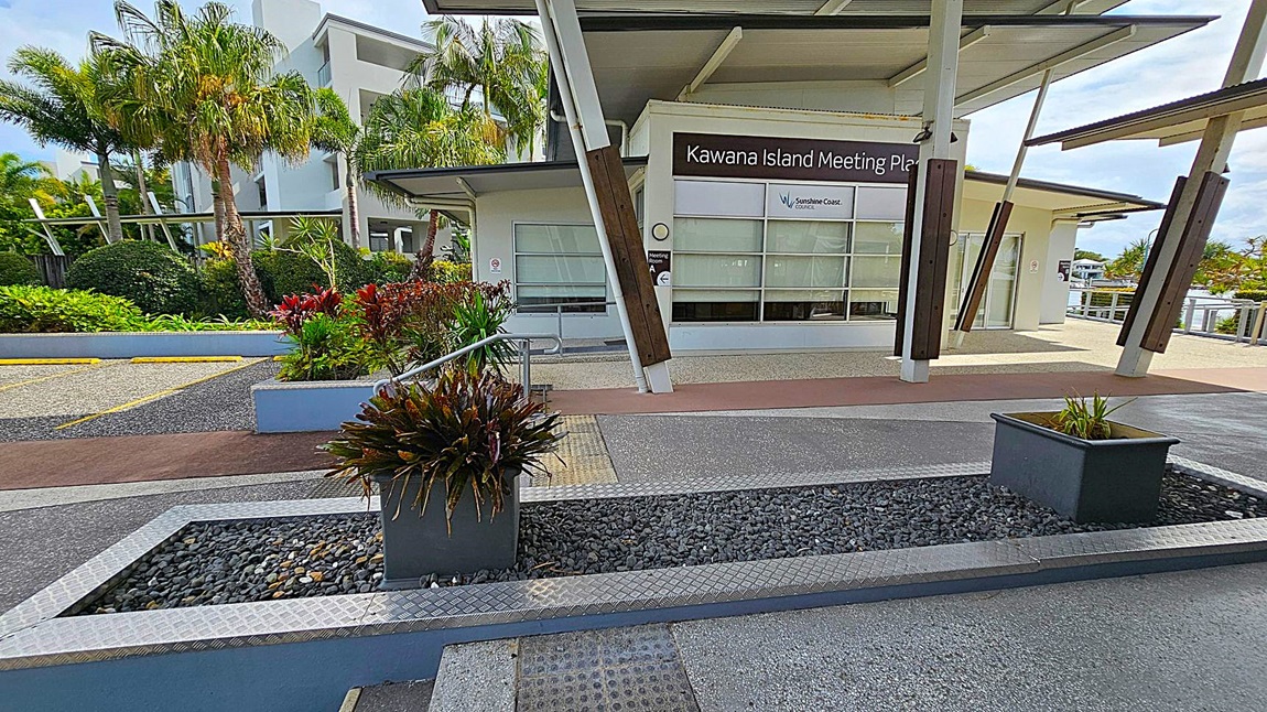 In the foreground, there are two rectangular planters with green plants, set on a bed of dark pebbles. The background features the Kawana Island Meeting Place building with large windows and a sign for the Sunshine Coast Council. To the left, there are lush green plants and palm trees. The ground is a mix of textured concrete and pebble surfaces, and the building has a modern design with angled supports and a flat roof. In the foreground, there are two rectangular planters with green plants, set on a bed of dark pebbles. The background features the Kawana Island Meeting Place building with large windows and a sign for the Sunshine Coast Council. To the left, there are lush green plants and palm trees. The ground is a mix of textured concrete and pebble surfaces, and the building has a modern design with angled supports and a flat roof.