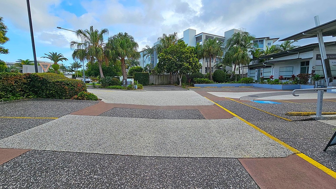 A street scene with a textured, multi-coloured pavement featuring grey, brown, and black sections. On the left, there are lush green bushes and palm trees, with a "Give Way" sign visible. In the centre, a modern white building is partially obscured by more palm trees and greenery. On the right, a building with a sloped roof and a sign is surrounded by landscaped plants. A metal railing is visible in the foreground. A street scene with a textured, multi-coloured pavement featuring grey, brown, and black sections. On the left, there are lush green bushes and palm trees, with a "Give Way" sign visible. In the centre, a modern white building is partially obscured by more palm trees and greenery. On the right, a building with a sloped roof and a sign is surrounded by landscaped plants. A metal railing is visible in the foreground.