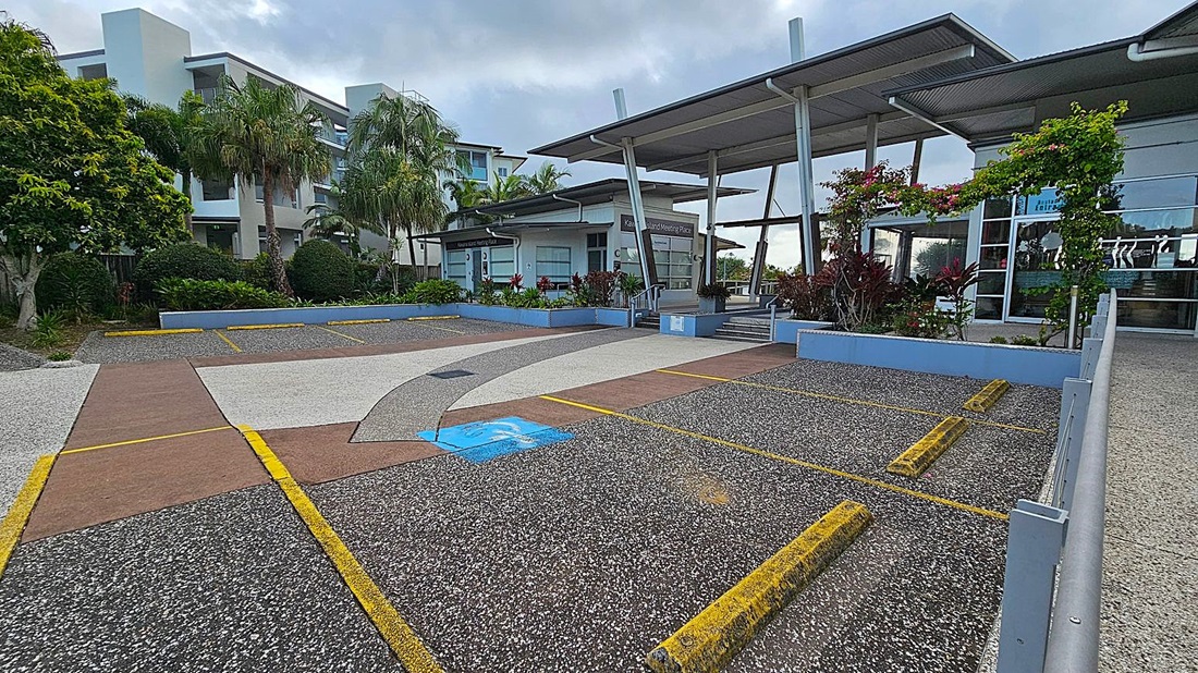 A modern building with a flat roof and large glass windows is on the right. In front, there are landscaped areas with shrubs and flowering plants. The foreground features a parking area with yellow lines and a designated blue disabled parking space. On the left, there are tall palm trees and a multi-storey building with balconies. The ground is a mix of textured concrete and asphalt. A modern building with a flat roof and large glass windows is on the right. In front, there are landscaped areas with shrubs and flowering plants. The foreground features a parking area with yellow lines and a designated blue disabled parking space. On the left, there are tall palm trees and a multi-storey building with balconies. The ground is a mix of textured concrete and asphalt.