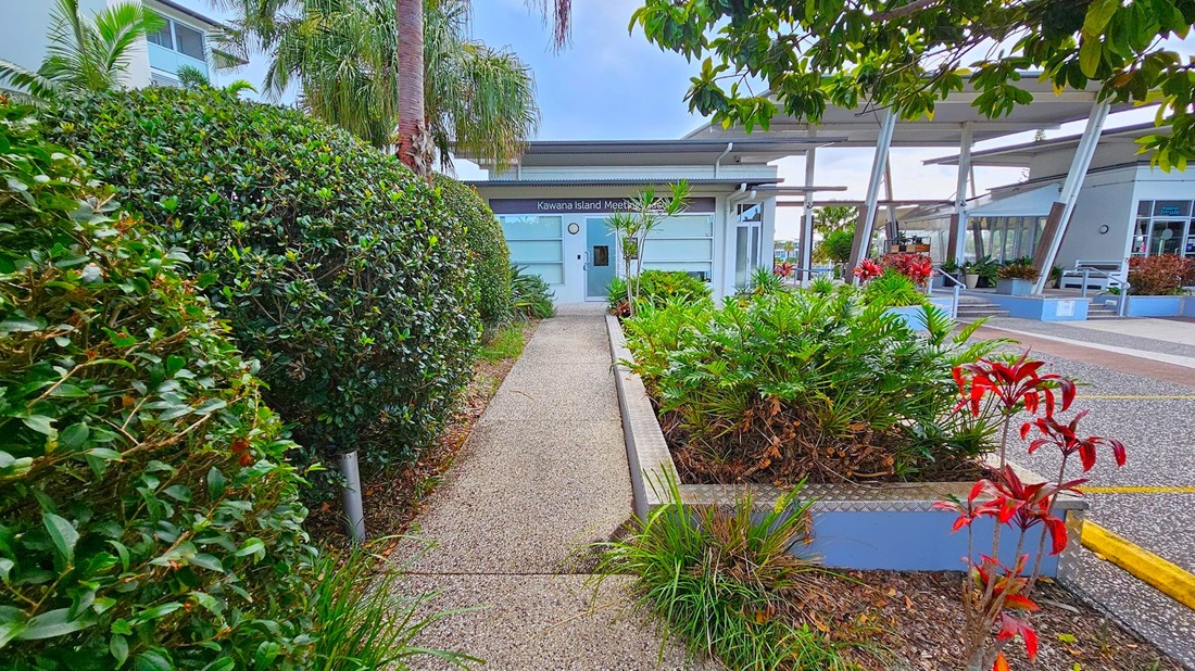 A pathway leads to a building with a sign reading "Kawana Island Meeting Place." On the left, dense green hedges and palm trees line the path. On the right, there are various plants, including ferns and red-leaved shrubs, in a raised garden bed. The building has large windows and a modern design. In the background, there are more structures with open seating areas and additional greenery. The ground is covered with a textured, speckled surface. A pathway leads to a building with a sign reading "Kawana Island Meeting Place." On the left, dense green hedges and palm trees line the path. On the right, there are various plants, including ferns and red-leaved shrubs, in a raised garden bed. The building has large windows and a modern design. In the background, there are more structures with open seating areas and additional greenery. The ground is covered with a textured, speckled surface.