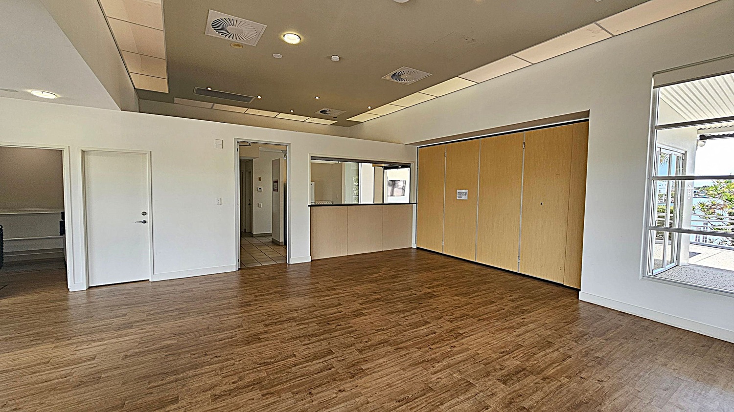 A room with wooden flooring and a light-coloured ceiling featuring recessed lighting and air vents. On the left, there are two white doors, one slightly ajar, leading to another area. In the centre, a counter with a window above it is set into the wall. On the right, large wooden sliding doors are closed, and a window reveals an outdoor view with greenery and a building. A room with wooden flooring and a light-coloured ceiling featuring recessed lighting and air vents. On the left, there are two white doors, one slightly ajar, leading to another area. In the centre, a counter with a window above it is set into the wall. On the right, large wooden sliding doors are closed, and a window reveals an outdoor view with greenery and a building.