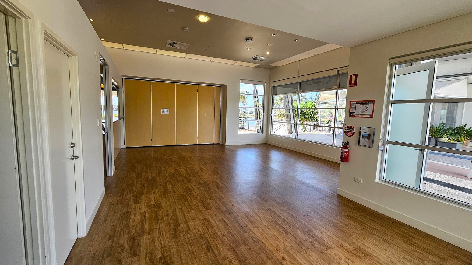 A spacious room with wooden flooring and a light-coloured ceiling featuring recessed lighting and air vents. On the left, there are two closed doors. The back wall has large windows and a set of wooden folding doors. On the right, more windows allow natural light in, with a fire extinguisher and safety instructions mounted on the wall. Outside, there are visible plants and a walkway. A spacious room with wooden flooring and a light-coloured ceiling featuring recessed lighting and air vents. On the left, there are two closed doors. The back wall has large windows and a set of wooden folding doors. On the right, more windows allow natural light in, with a fire extinguisher and safety instructions mounted on the wall. Outside, there are visible plants and a walkway.