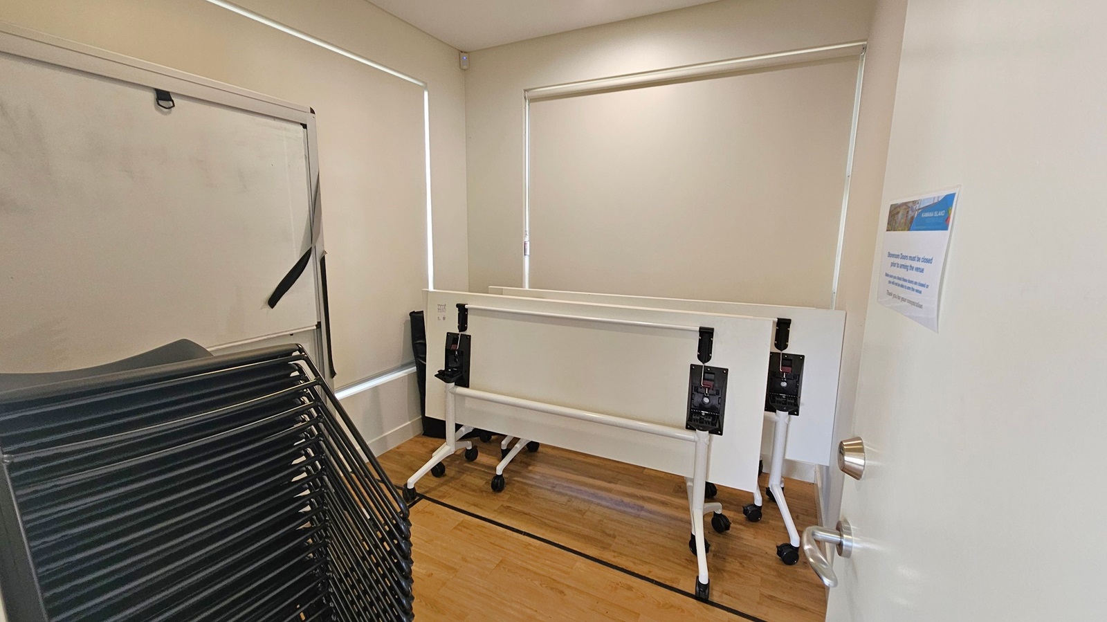 A small room with light-coloured walls and wooden flooring. On the left, there is a stack of black chairs. In the centre, two white folding tables with wheels are positioned against the wall. The windows have closed blinds. On the right, a white door is partially open, with a silver handle and a notice attached to it. A small room with light-coloured walls and wooden flooring. On the left, there is a stack of black chairs. In the centre, two white folding tables with wheels are positioned against the wall. The windows have closed blinds. On the right, a white door is partially open, with a silver handle and a notice attached to it.