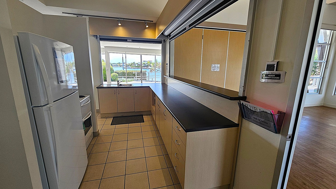 A kitchen with a tiled floor and a black countertop. On the left, there's a white refrigerator and a stove. The countertop extends along the right side with multiple drawers. In the background, large windows offer a view of a waterfront with trees and buildings. A small mat is placed near the sink. A wall-mounted security system and a holder with papers marked "Out of Order" are visible on the right. The ceiling has recessed lighting. A kitchen with a tiled floor and a black countertop. On the left, there's a white refrigerator and a stove. The countertop extends along the right side with multiple drawers. In the background, large windows offer a view of a waterfront with trees and buildings. A small mat is placed near the sink. A wall-mounted security system and a holder with papers marked "Out of Order" are visible on the right. The ceiling has recessed lighting.
