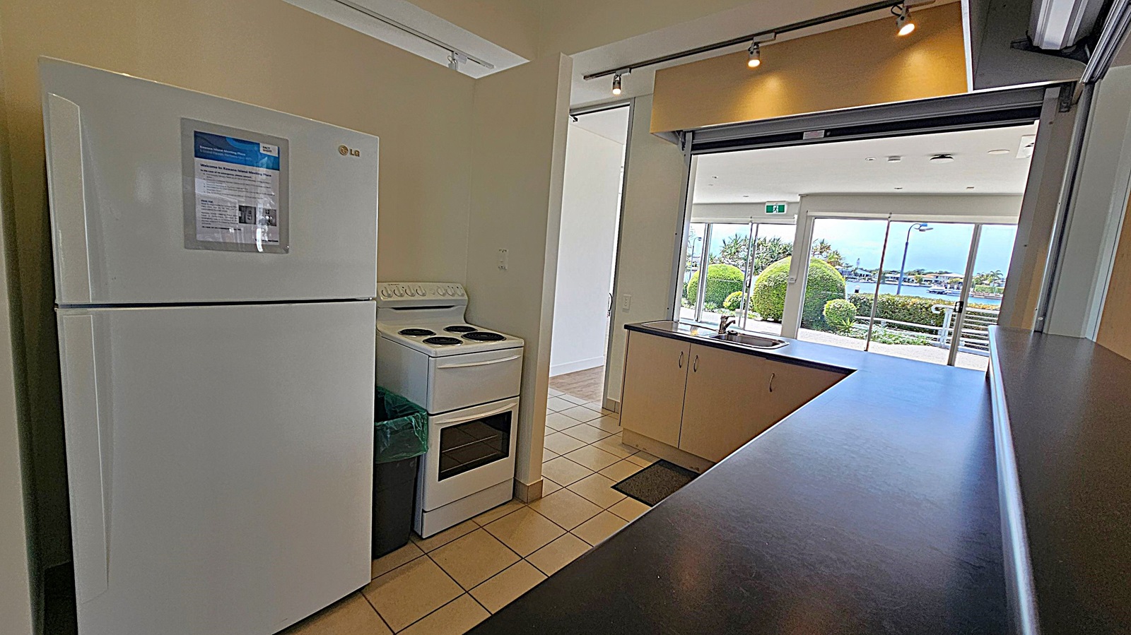 A kitchen area with a white fridge on the left, featuring a notice on its door. Next to it is a white electric stove. The floor is tiled. On the right, a counter with a sink overlooks a large window. Beyond the window, there are neatly trimmed bushes and a view of water and distant buildings. The ceiling has track lighting. A kitchen area with a white fridge on the left, featuring a notice on its door. Next to it is a white electric stove. The floor is tiled. On the right, a counter with a sink overlooks a large window. Beyond the window, there are neatly trimmed bushes and a view of water and distant buildings. The ceiling has track lighting.