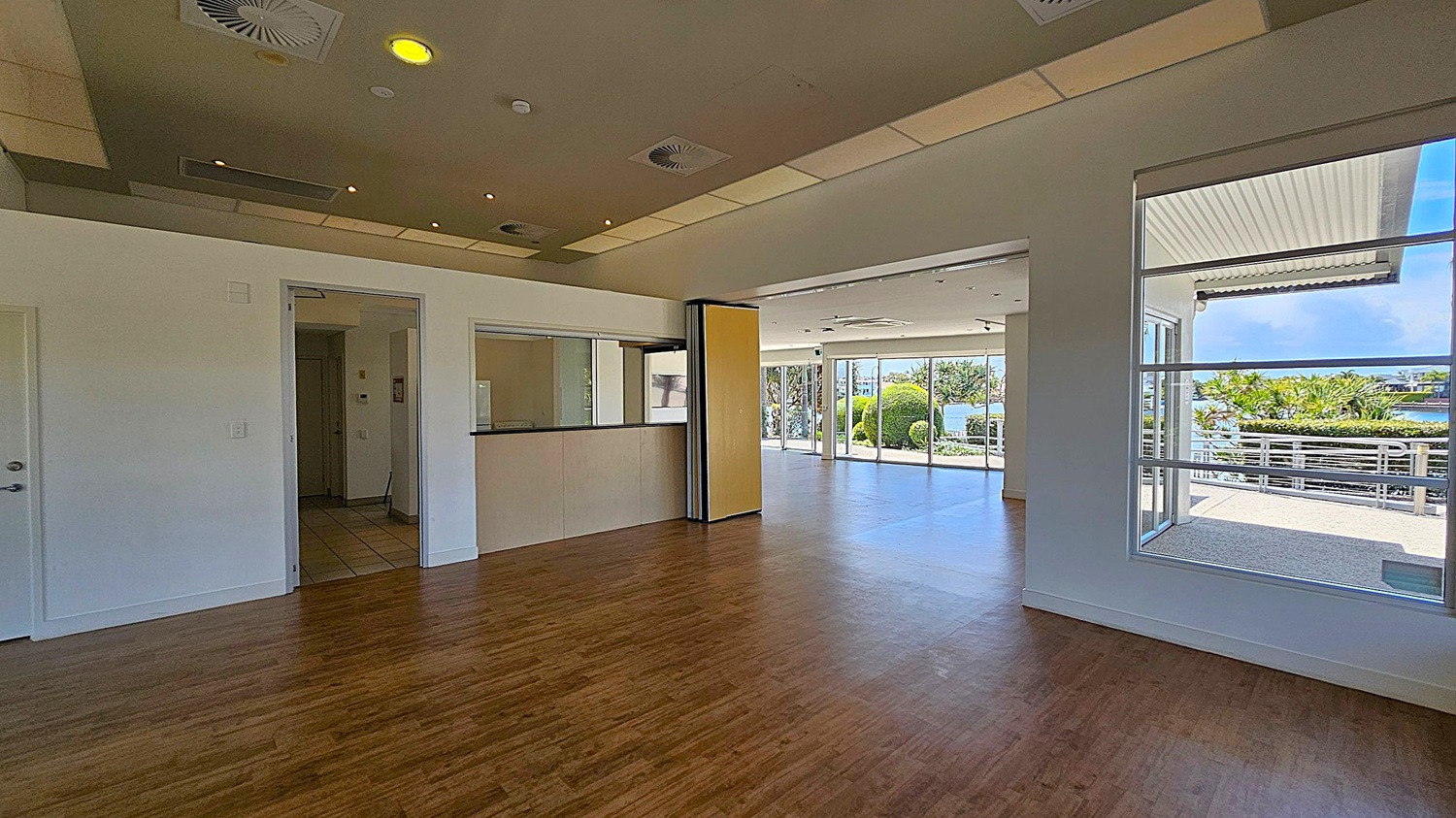 A spacious room with wooden flooring and a ceiling featuring recessed lighting and air vents. On the left, there is a small counter with a window and a doorway leading to another room. The right side has large windows and glass doors opening to an outdoor area with greenery and a view of water. The outdoor area has a railing and a covered section. A spacious room with wooden flooring and a ceiling featuring recessed lighting and air vents. On the left, there is a small counter with a window and a doorway leading to another room. The right side has large windows and glass doors opening to an outdoor area with greenery and a view of water. The outdoor area has a railing and a covered section.