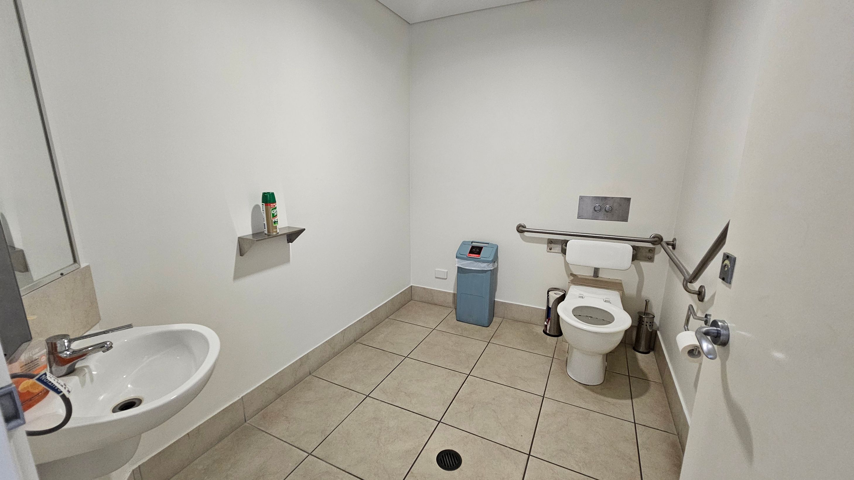A bathroom with beige tiled flooring and white walls. On the left, there's a white sink with a silver tap and a mirror above it. A small shelf holds a can of spray. In the centre, a blue bin is placed against the wall. On the right, a toilet with a backrest is equipped with a grab rail. A toilet roll holder is mounted on the wall, and a small bin is nearby. The ceiling is plain white. A bathroom with beige tiled flooring and white walls. On the left, there's a white sink with a silver tap and a mirror above it. A small shelf holds a can of spray. In the centre, a blue bin is placed against the wall. On the right, a toilet with a backrest is equipped with a grab rail. A toilet roll holder is mounted on the wall, and a small bin is nearby. The ceiling is plain white.