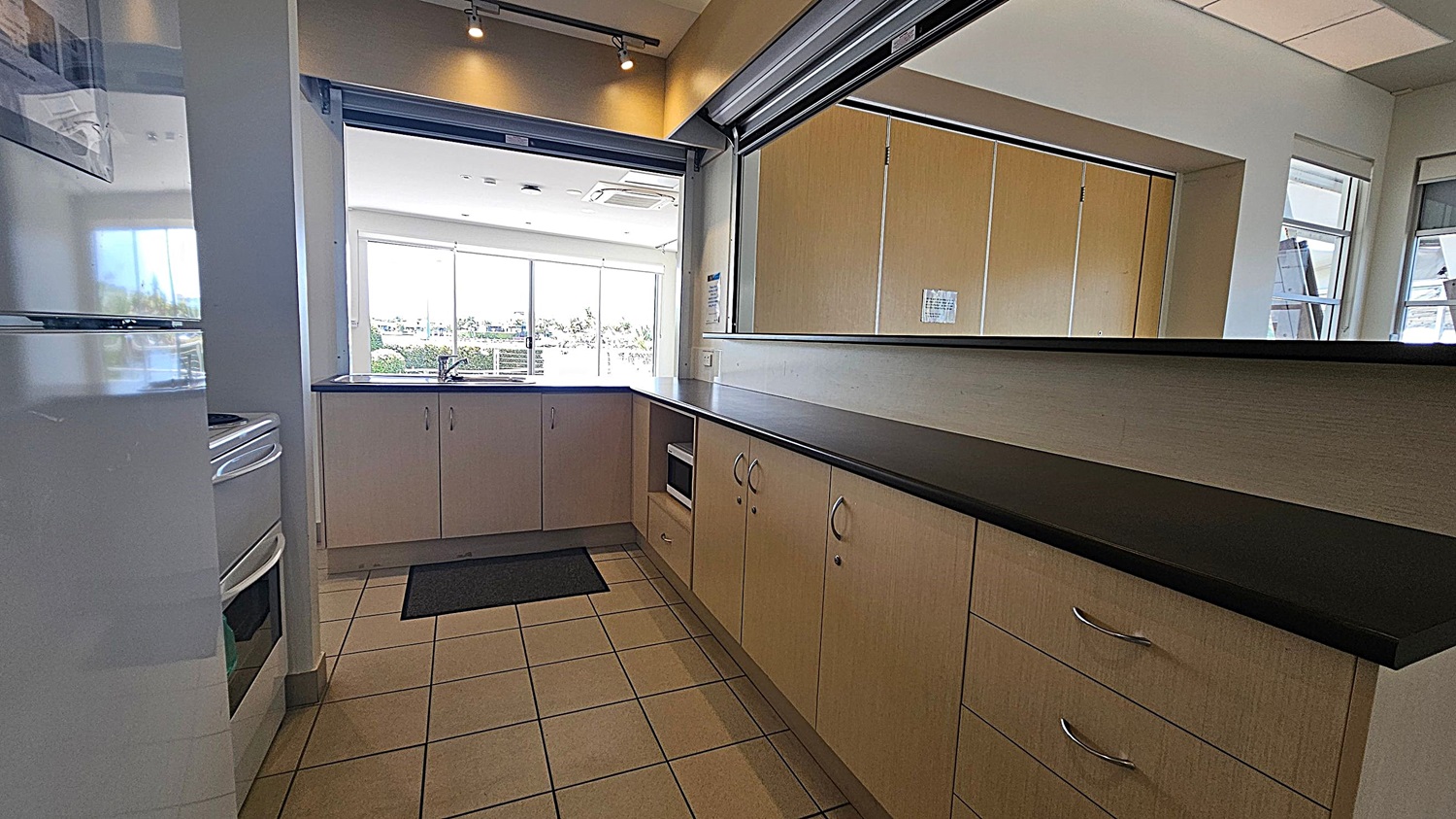 A kitchen area with a stainless steel sink and drainer on the left, set into a dark countertop. Below the counter are light-coloured cabinets and a microwave. On the right, a white wall features an Automated External Defibrillator (AED) in a wall-mounted case. In the background, large windows and glass doors reveal an outdoor area with plants and a pathway. The floor is tiled, and the ceiling is not visible. A kitchen area with a stainless steel sink and drainer on the left, set into a dark countertop. Below the counter are light-coloured cabinets and a microwave. On the right, a white wall features an Automated External Defibrillator (AED) in a wall-mounted case. In the background, large windows and glass doors reveal an outdoor area with plants and a pathway. The floor is tiled, and the ceiling is not visible.
