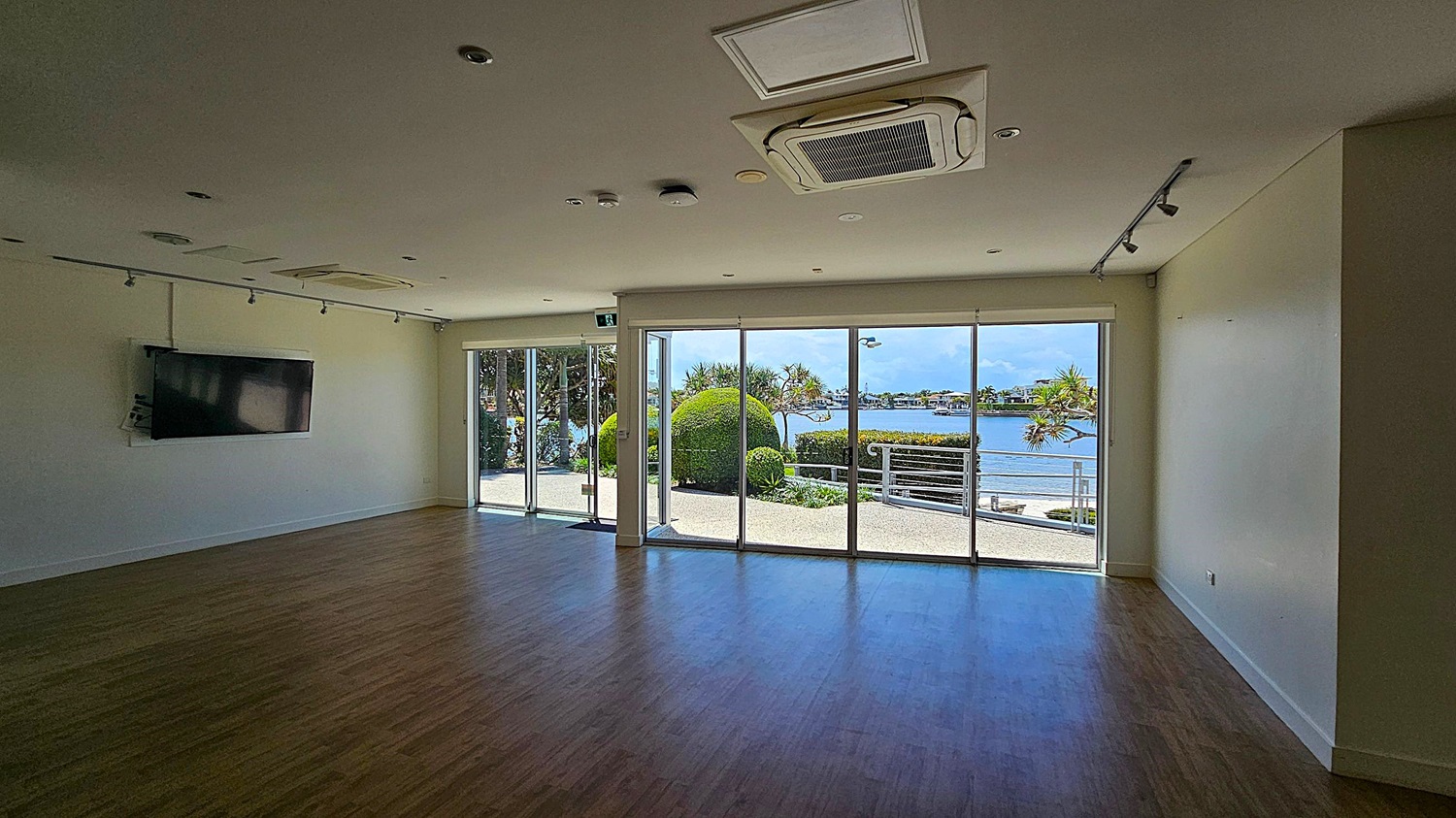 A spacious room with wooden flooring and a white ceiling featuring recessed lighting and an air conditioning unit. On the left wall, a flat-screen TV is mounted. The right side has large glass sliding doors leading to an outdoor area with neatly trimmed bushes and a view of a body of water and distant buildings. The room is well-lit by natural light coming through the glass doors. A spacious room with wooden flooring and a white ceiling featuring recessed lighting and an air conditioning unit. On the left wall, a flat-screen TV is mounted. The right side has large glass sliding doors leading to an outdoor area with neatly trimmed bushes and a view of a body of water and distant buildings. The room is well-lit by natural light coming through the glass doors.