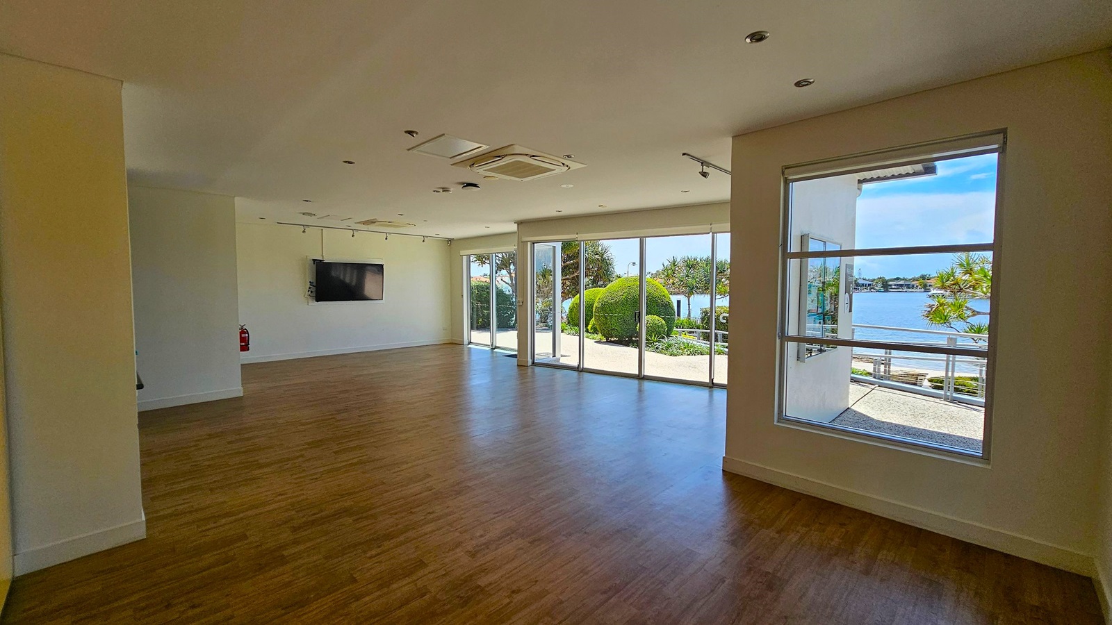 A spacious room with wooden flooring and a white ceiling featuring recessed lights and an air conditioning unit. On the left, a wall-mounted television and a fire extinguisher are visible. The right side has large windows and glass doors, offering a view of a garden with neatly trimmed bushes and a body of water in the background. The room is well-lit with natural light streaming in through the windows. A spacious room with wooden flooring and a white ceiling featuring recessed lights and an air conditioning unit. On the left, a wall-mounted television and a fire extinguisher are visible. The right side has large windows and glass doors, offering a view of a garden with neatly trimmed bushes and a body of water in the background. The room is well-lit with natural light streaming in through the windows.