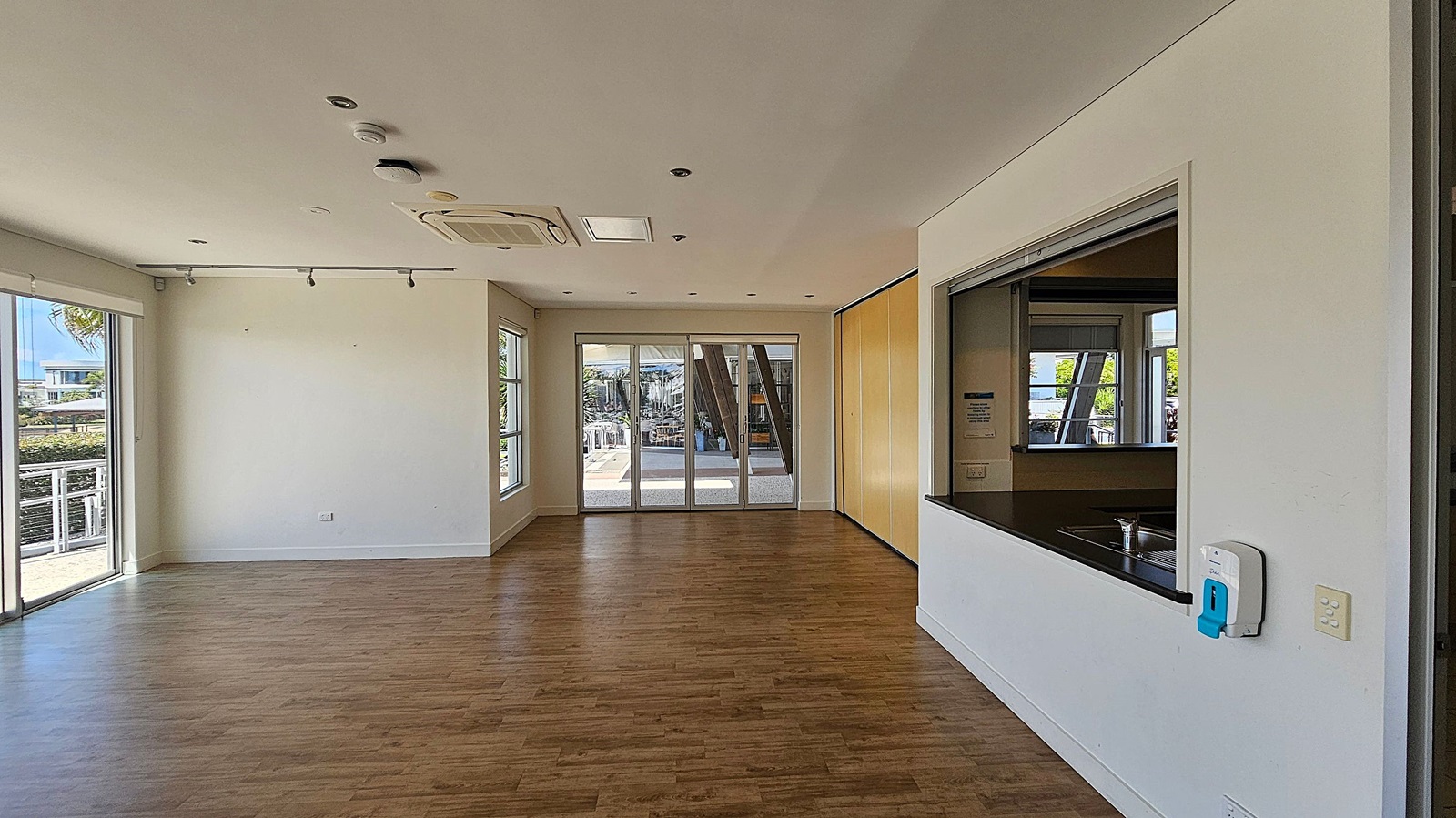 A spacious room with wooden flooring and white walls. On the left, large windows and a sliding glass door provide natural light and a view outside. In the centre, a set of glass doors leads to an outdoor area. On the right, there's a serving hatch with a countertop and a sink, and a wall-mounted hand sanitiser dispenser. The ceiling is fitted with recessed lights and an air conditioning unit. A spacious room with wooden flooring and white walls. On the left, large windows and a sliding glass door provide natural light and a view outside. In the centre, a set of glass doors leads to an outdoor area. On the right, there's a serving hatch with a countertop and a sink, and a wall-mounted hand sanitiser dispenser. The ceiling is fitted with recessed lights and an air conditioning unit.