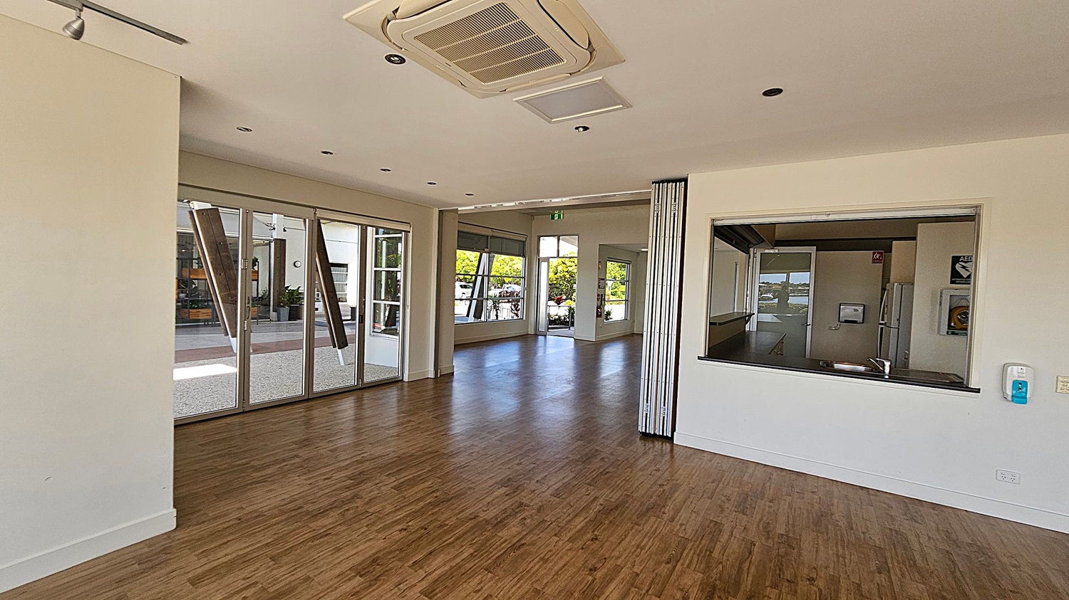 A spacious room with wooden flooring and a white ceiling featuring recessed lights and an air conditioning unit. On the left, there are large glass doors leading outside. In the centre, a hallway with more windows and an open door reveals greenery outside. On the right, a large serving window opens into a kitchen area, with a sink visible. A hand sanitiser dispenser is mounted on the wall near the window. A spacious room with wooden flooring and a white ceiling featuring recessed lights and an air conditioning unit. On the left, there are large glass doors leading outside. In the centre, a hallway with more windows and an open door reveals greenery outside. On the right, a large serving window opens into a kitchen area, with a sink visible. A hand sanitiser dispenser is mounted on the wall near the window.