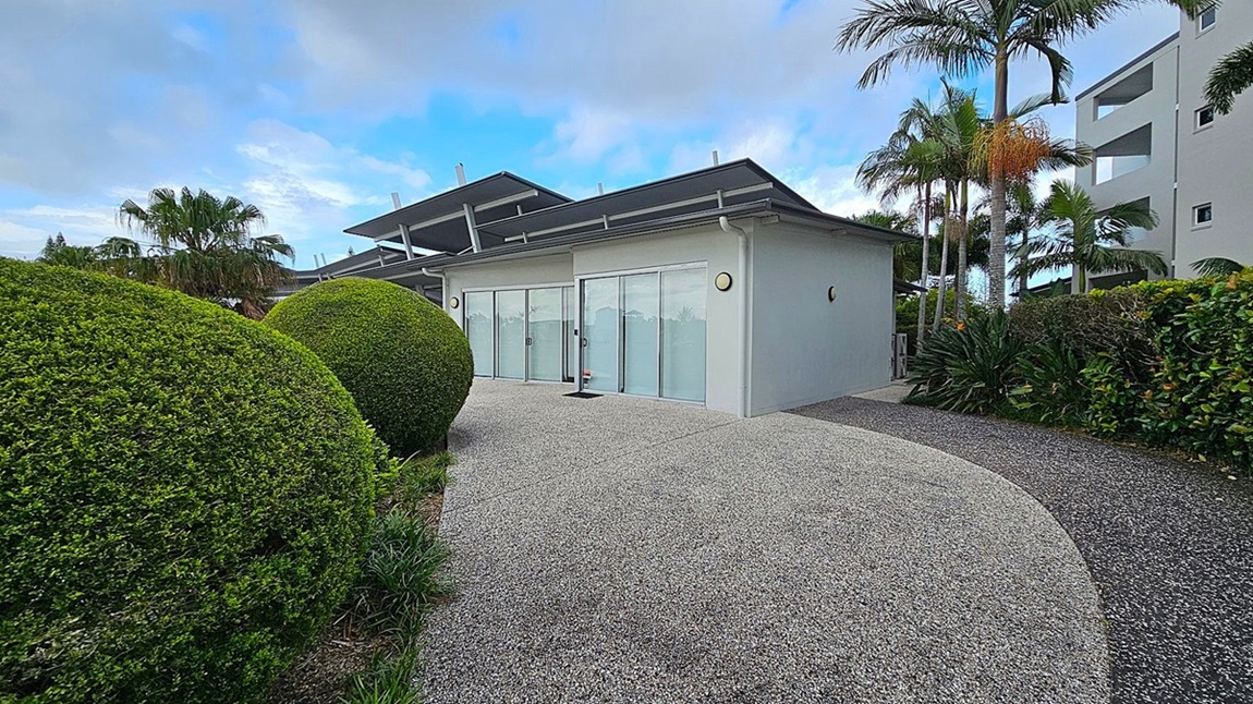 A modern building with large glass sliding doors is in the centre, surrounded by a neatly landscaped garden. On the left, there are large, round, manicured bushes. On the right, tall palm trees and dense greenery line the path. The ground is covered with a speckled grey concrete path that curves around the building. The building has a flat roof with angular extensions. A modern building with large glass sliding doors is in the centre, surrounded by a neatly landscaped garden. On the left, there are large, round, manicured bushes. On the right, tall palm trees and dense greenery line the path. The ground is covered with a speckled grey concrete path that curves around the building. The building has a flat roof with angular extensions.