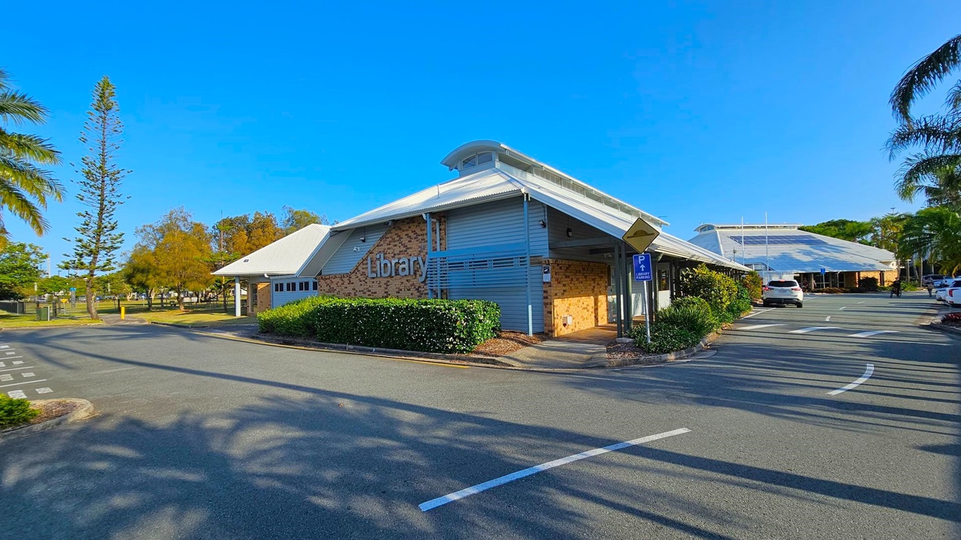 A library building with a modern design, featuring a sloped roof and a mix of brick and grey panelling. The word "Library" is prominently displayed on the side. In the foreground, there is a paved road with parking spaces and a sign indicating library parking. On the left, tall trees and a grassy area are visible. On the right, more parking spaces and a few cars are present, with palm trees lining the area. Shadows of trees are cast on the pavement. A library building with a modern design, featuring a sloped roof and a mix of brick and grey panelling. The word "Library" is prominently displayed on the side. In the foreground, there is a paved road with parking spaces and a sign indicating library parking. On the left, tall trees and a grassy area are visible. On the right, more parking spaces and a few cars are present, with palm trees lining the area. Shadows of trees are cast on the pavement.