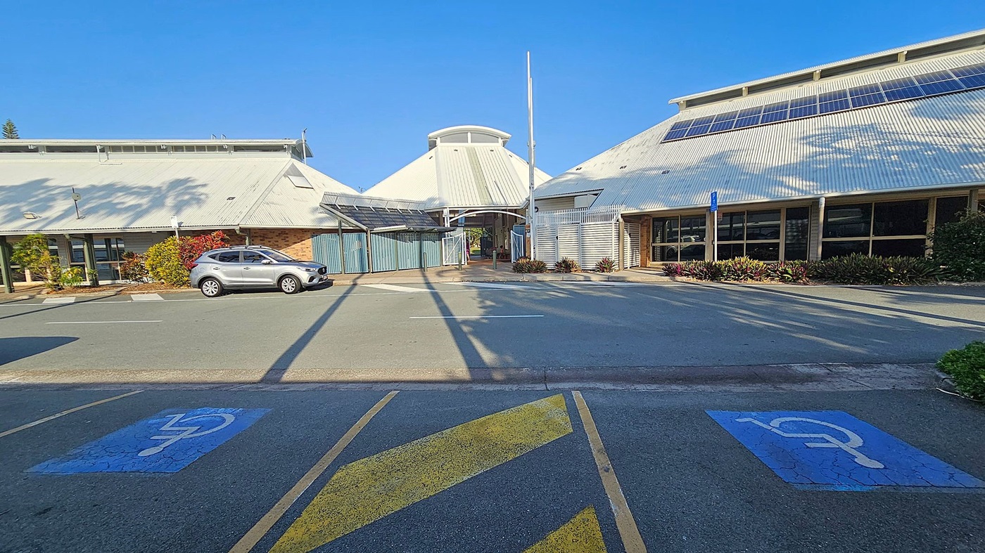 A car park with two marked disabled parking spaces in the foreground. A silver car is parked on the left side near a building with a sloped roof. The building has solar panels on the roof and is surrounded by plants. In the centre, there is a covered walkway leading to an entrance. The right side features another building with large windows and more solar panels. Shadows of trees are cast on the ground and buildings. A car park with two marked disabled parking spaces in the foreground. A silver car is parked on the left side near a building with a sloped roof. The building has solar panels on the roof and is surrounded by plants. In the centre, there is a covered walkway leading to an entrance. The right side features another building with large windows and more solar panels. Shadows of trees are cast on the ground and buildings.