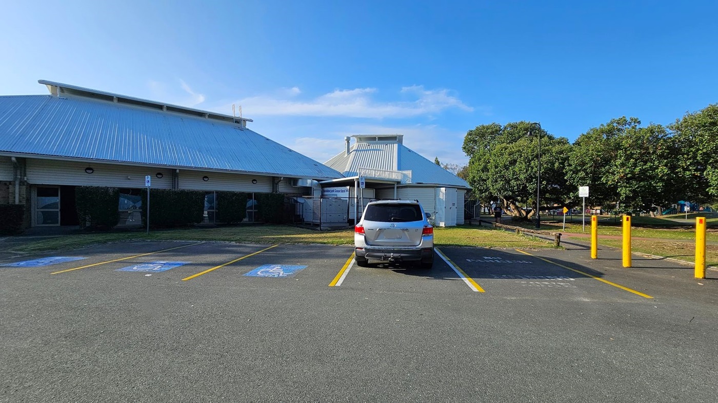 A car park with a silver SUV parked in a designated disabled parking space. The ground is marked with blue and white symbols. To the left, a large building with a corrugated metal roof and brick walls. On the right, yellow bollards line the edge of the parking area. In the background, there are trees and a grassy area with a path. A car park with a silver SUV parked in a designated disabled parking space. The ground is marked with blue and white symbols. To the left, a large building with a corrugated metal roof and brick walls. On the right, yellow bollards line the edge of the parking area. In the background, there are trees and a grassy area with a path.