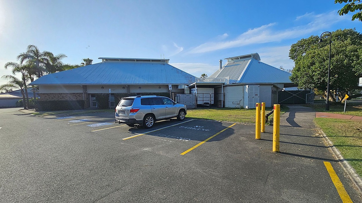 A car park with a silver SUV parked in a designated space on the left. The ground is asphalt with yellow parking lines. In the centre, a building with a metal roof and brick walls is visible. On the right, there are three yellow bollards and a pathway leading to a grassy area with a large tree. A lamppost and a sign are near the tree. Palm trees are in the background on the left. A car park with a silver SUV parked in a designated space on the left. The ground is asphalt with yellow parking lines. In the centre, a building with a metal roof and brick walls is visible. On the right, there are three yellow bollards and a pathway leading to a grassy area with a large tree. A lamppost and a sign are near the tree. Palm trees are in the background on the left.