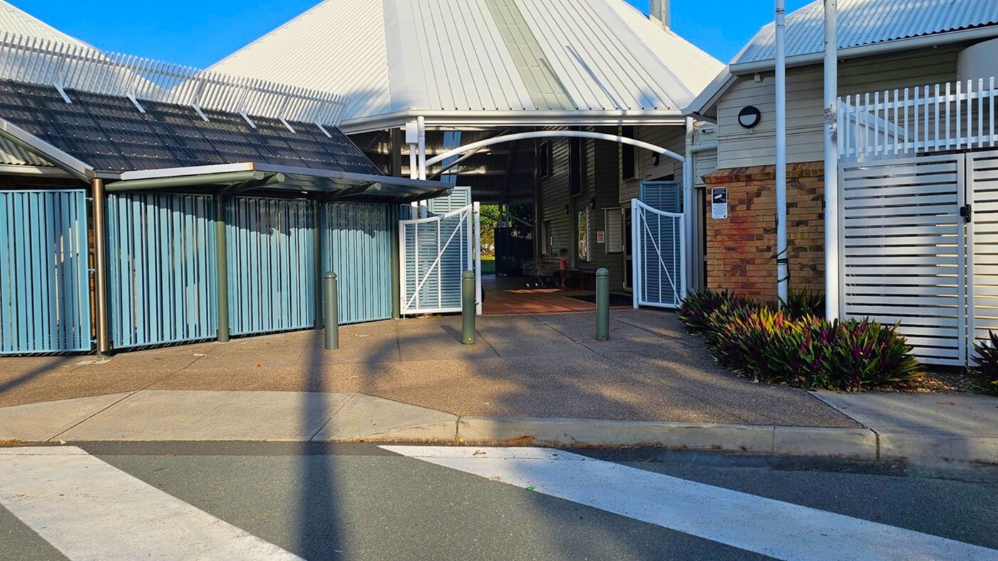 A building entrance with a white arched gate in the centre, flanked by blue vertical slatted panels on the left and a brick wall with white horizontal slats on the right. The roof is corrugated metal. There are three bollards in front of the entrance. The ground is paved, and there are plants on the right side. A pedestrian crossing is visible in the foreground. A building entrance with a white arched gate in the centre, flanked by blue vertical slatted panels on the left and a brick wall with white horizontal slats on the right. The roof is corrugated metal. There are three bollards in front of the entrance. The ground is paved, and there are plants on the right side. A pedestrian crossing is visible in the foreground.