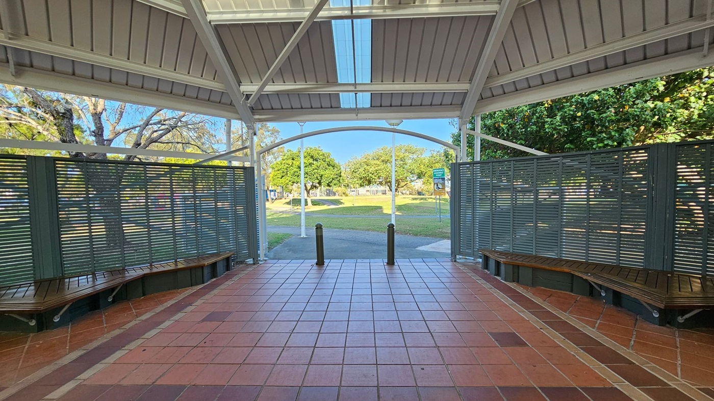 A covered outdoor area with a tiled floor and a metal roof. On the left and right, there are green slatted partitions with wooden benches attached. In the background, there is a grassy park with trees and a pathway. Two bollards are positioned at the entrance to the park. A covered outdoor area with a tiled floor and a metal roof. On the left and right, there are green slatted partitions with wooden benches attached. In the background, there is a grassy park with trees and a pathway. Two bollards are positioned at the entrance to the park.