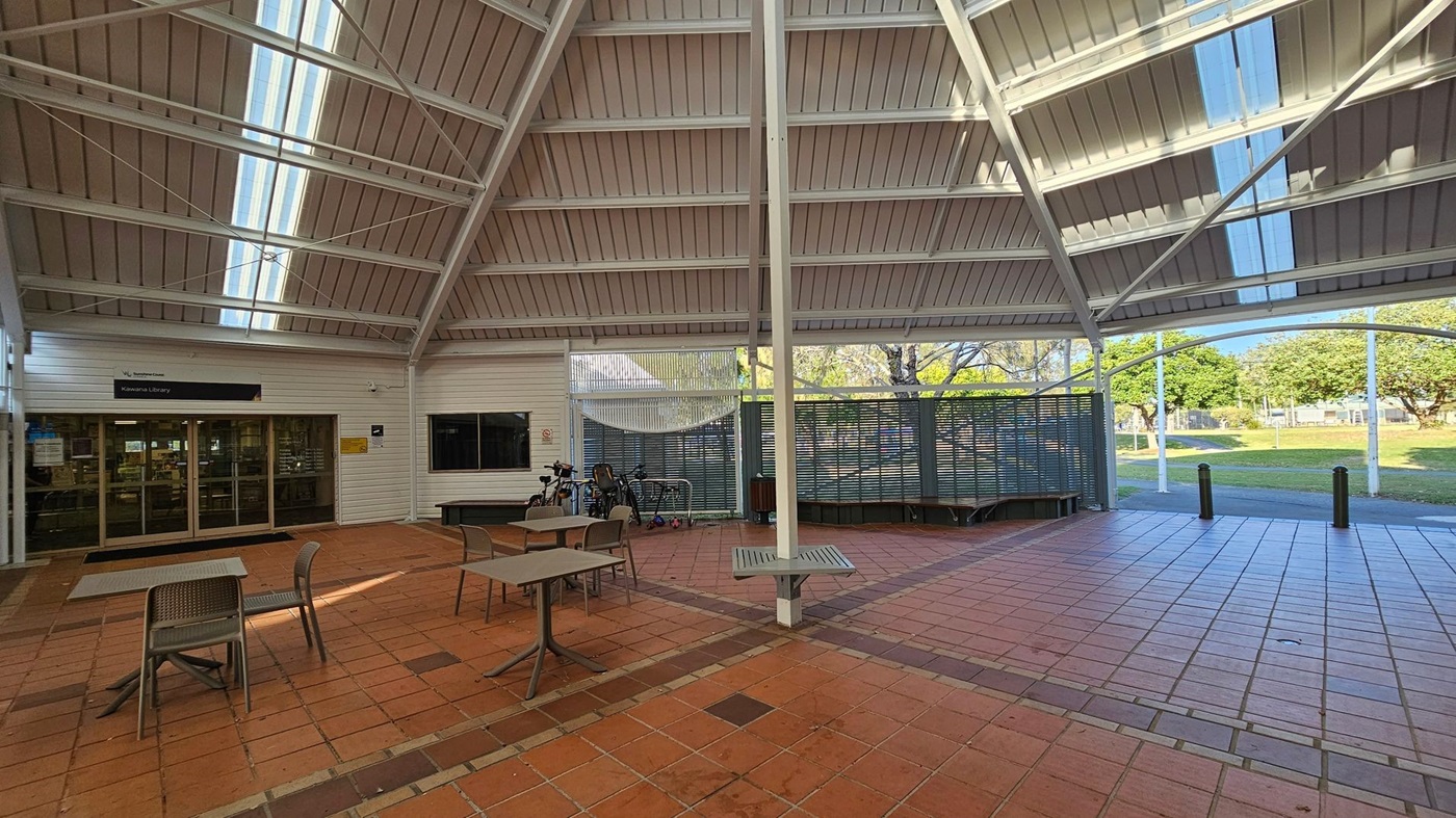 A covered outdoor area with a high, slanted roof featuring skylights. On the left, there is an entrance to a library with glass doors. In front, several tables and chairs are placed on a tiled floor. Bicycles are parked near a metal fence in the background. The right side opens to a grassy area with trees visible through the open space. The ceiling is supported by white beams. A covered outdoor area with a high, slanted roof featuring skylights. On the left, there is an entrance to a library with glass doors. In front, several tables and chairs are placed on a tiled floor. Bicycles are parked near a metal fence in the background. The right side opens to a grassy area with trees visible through the open space. The ceiling is supported by white beams.