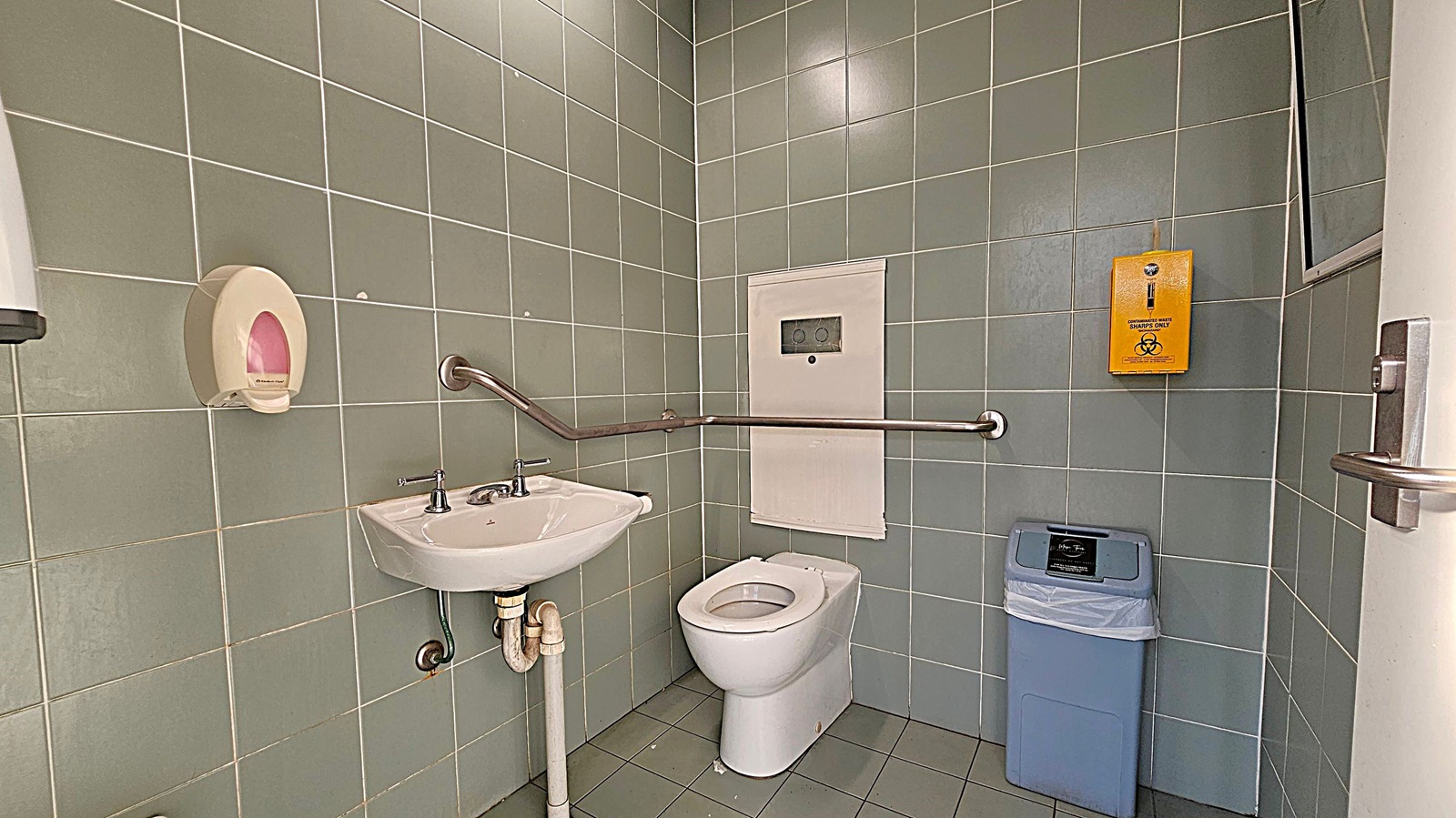 A bathroom with green tiled walls and floor. On the left, there's a white sink with a metal tap and a soap dispenser above it. A metal grab bar runs along the wall. In the centre, a white toilet is positioned beneath a panel. On the right, a yellow sharps disposal box is mounted on the wall above a blue bin. A door with a metal handle is partially visible on the far right. A bathroom with green tiled walls and floor. On the left, there's a white sink with a metal tap and a soap dispenser above it. A metal grab bar runs along the wall. In the centre, a white toilet is positioned beneath a panel. On the right, a yellow sharps disposal box is mounted on the wall above a blue bin. A door with a metal handle is partially visible on the far right.