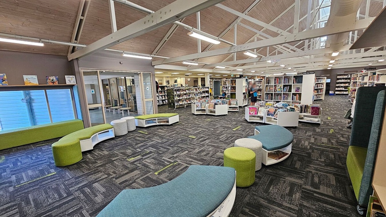 A spacious library interior with a high, wooden ceiling and exposed beams. The floor is covered with patterned carpet tiles in grey and green. On the left, there are green and grey curved benches near a large window. In the centre, there are white bookshelves filled with books, and a person is standing nearby. The right side has more bookshelves and seating areas. The background features a glass-walled room and additional bookshelves. A spacious library interior with a high, wooden ceiling and exposed beams. The floor is covered with patterned carpet tiles in grey and green. On the left, there are green and grey curved benches near a large window. In the centre, there are white bookshelves filled with books, and a person is standing nearby. The right side has more bookshelves and seating areas. The background features a glass-walled room and additional bookshelves.