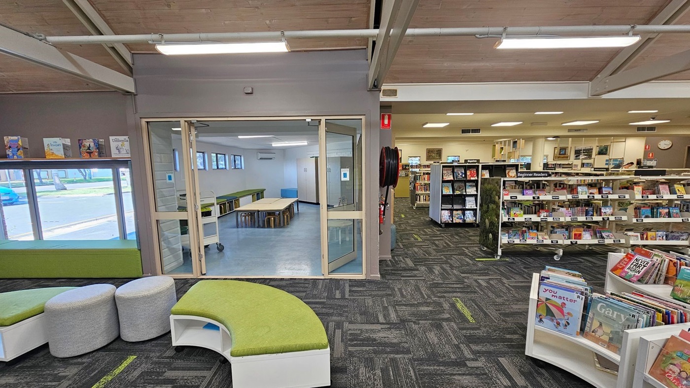 A library interior with a grey and green carpeted floor. On the left, there are green cushioned benches and round grey stools. A glass door in the centre leads to a room with tables and stools, featuring a light blue floor. On the right, there are bookshelves labelled "Beginner Readers" filled with books. The ceiling is wooden with exposed beams and fluorescent lights. A window on the left shows a view of the outside. A library interior with a grey and green carpeted floor. On the left, there are green cushioned benches and round grey stools. A glass door in the centre leads to a room with tables and stools, featuring a light blue floor. On the right, there are bookshelves labelled "Beginner Readers" filled with books. The ceiling is wooden with exposed beams and fluorescent lights. A window on the left shows a view of the outside.