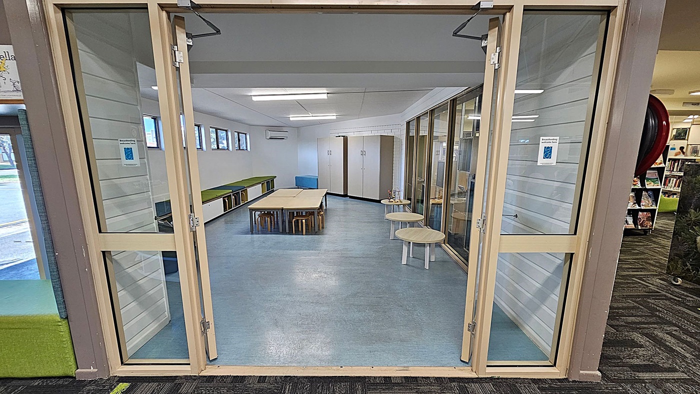 A room with glass doors and a blue floor. Inside, there are tables and stools in the centre, with green cushioned benches along the left wall. On the right, there are small round tables and a glass partition. The back wall has storage cabinets and small windows near the ceiling. The ceiling is white with fluorescent lights. The area outside the room has a patterned carpet with green and black designs. A room with glass doors and a blue floor. Inside, there are tables and stools in the centre, with green cushioned benches along the left wall. On the right, there are small round tables and a glass partition. The back wall has storage cabinets and small windows near the ceiling. The ceiling is white with fluorescent lights. The area outside the room has a patterned carpet with green and black designs.