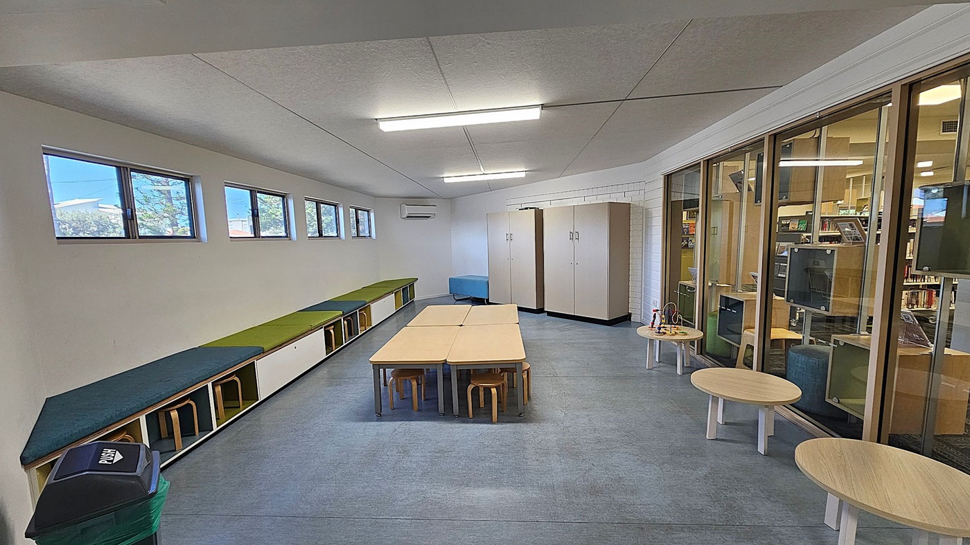 A room with a row of small windows on the left wall, above a long bench with green and blue cushions. Below the bench are stools. In the centre, there are light-coloured tables with small wooden stools. The right side features large glass panels showing another room with bookshelves. Near the glass panels are round wooden tables with toys. The floor is grey, and the ceiling has fluorescent lights. A bin is in the foreground on the left. A room with a row of small windows on the left wall, above a long bench with green and blue cushions. Below the bench are stools. In the centre, there are light-coloured tables with small wooden stools. The right side features large glass panels showing another room with bookshelves. Near the glass panels are round wooden tables with toys. The floor is grey, and the ceiling has fluorescent lights. A bin is in the foreground on the left.