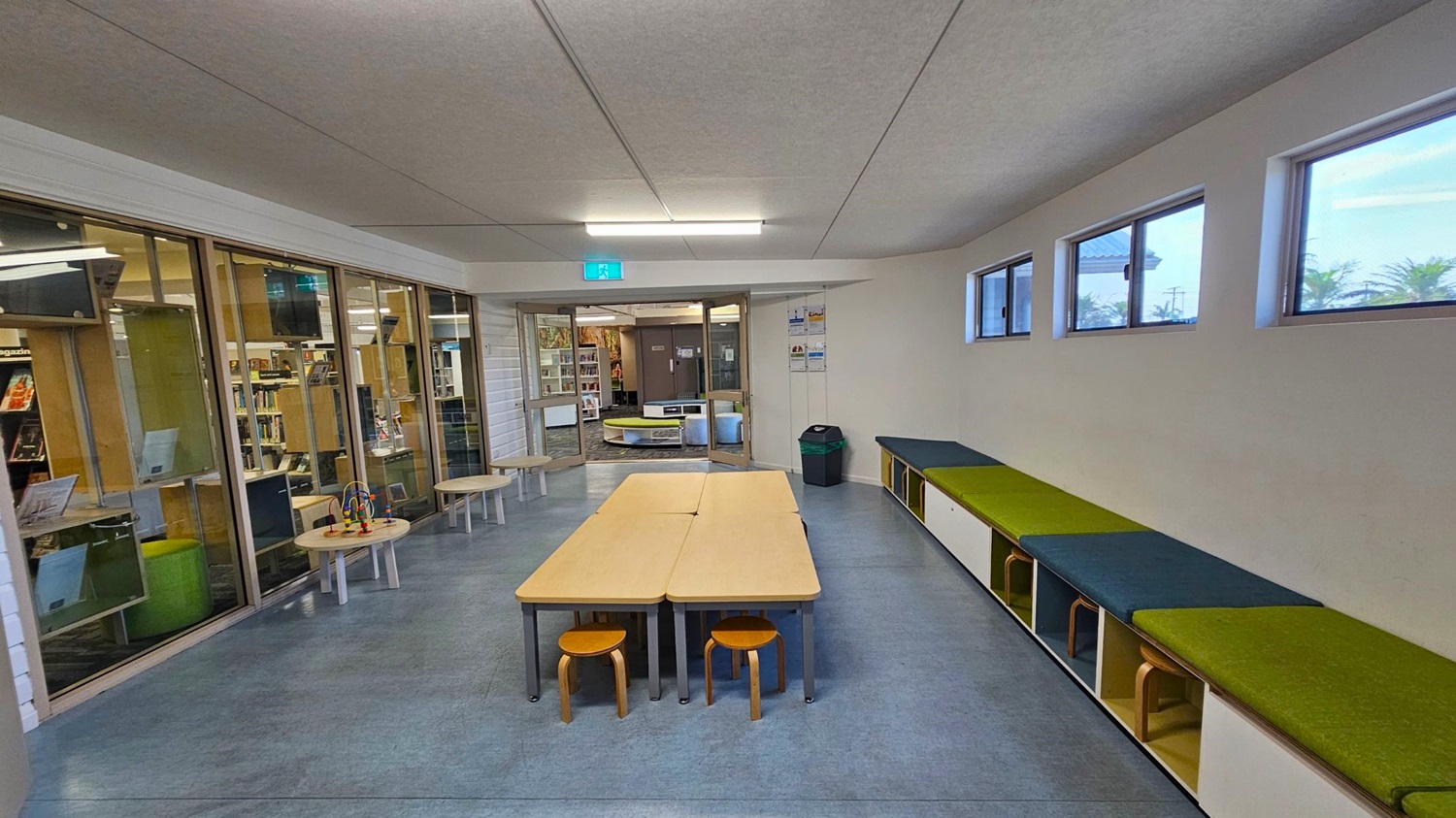 A room with a grey ceiling and blue-grey floor. On the left, there are glass walls with a view into a library, featuring bookshelves and small round tables with toys. In the centre, two rectangular tables are pushed together with small wooden stools underneath. On the right, a long bench with green and blue cushions sits below a row of small windows. A green and black bin is near the entrance, which leads to another room with more bookshelves. A room with a grey ceiling and blue-grey floor. On the left, there are glass walls with a view into a library, featuring bookshelves and small round tables with toys. In the centre, two rectangular tables are pushed together with small wooden stools underneath. On the right, a long bench with green and blue cushions sits below a row of small windows. A green and black bin is near the entrance, which leads to another room with more bookshelves.