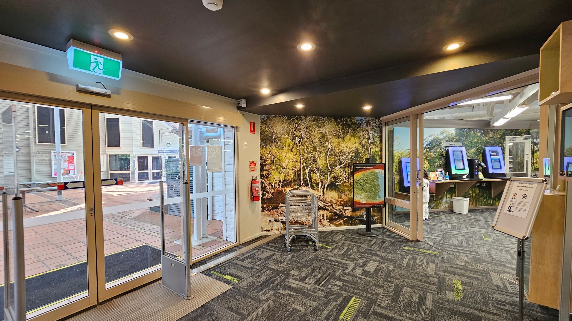 Glass entrance doors to a library with a "Zero Tolerance" sign on the left and opening hours on the right. Inside, bookshelves are visible with books and displays. The floor is tiled outside and carpeted inside. The ceiling has recessed lighting. Glass entrance doors to a library with a "Zero Tolerance" sign on the left and opening hours on the right. Inside, bookshelves are visible with books and displays. The floor is tiled outside and carpeted inside. The ceiling has recessed lighting.