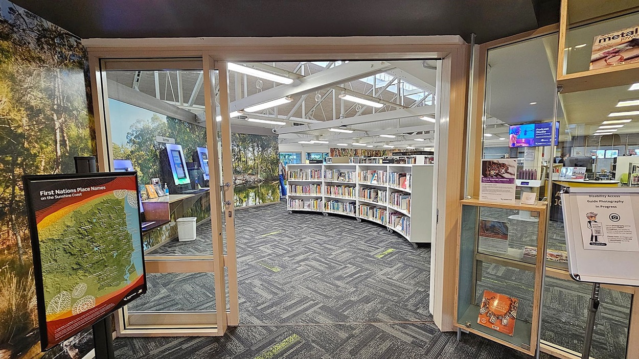 A library entrance with glass doors. On the left, a sign displays "First Nations Place Names on the Sunshine Coast" with a map. Behind it, a wall features a nature mural. Inside, curved bookshelves are filled with books. On the right, a glass display case holds informational materials, and a sign mentions "Disability Access Guide Photography in Progress." The floor is carpeted with a grey and green pattern, and the ceiling has exposed white beams with fluorescent lights. A library entrance with glass doors. On the left, a sign displays "First Nations Place Names on the Sunshine Coast" with a map. Behind it, a wall features a nature mural. Inside, curved bookshelves are filled with books. On the right, a glass display case holds informational materials, and a sign mentions "Disability Access Guide Photography in Progress." The floor is carpeted with a grey and green pattern, and the ceiling has exposed white beams with fluorescent lights.