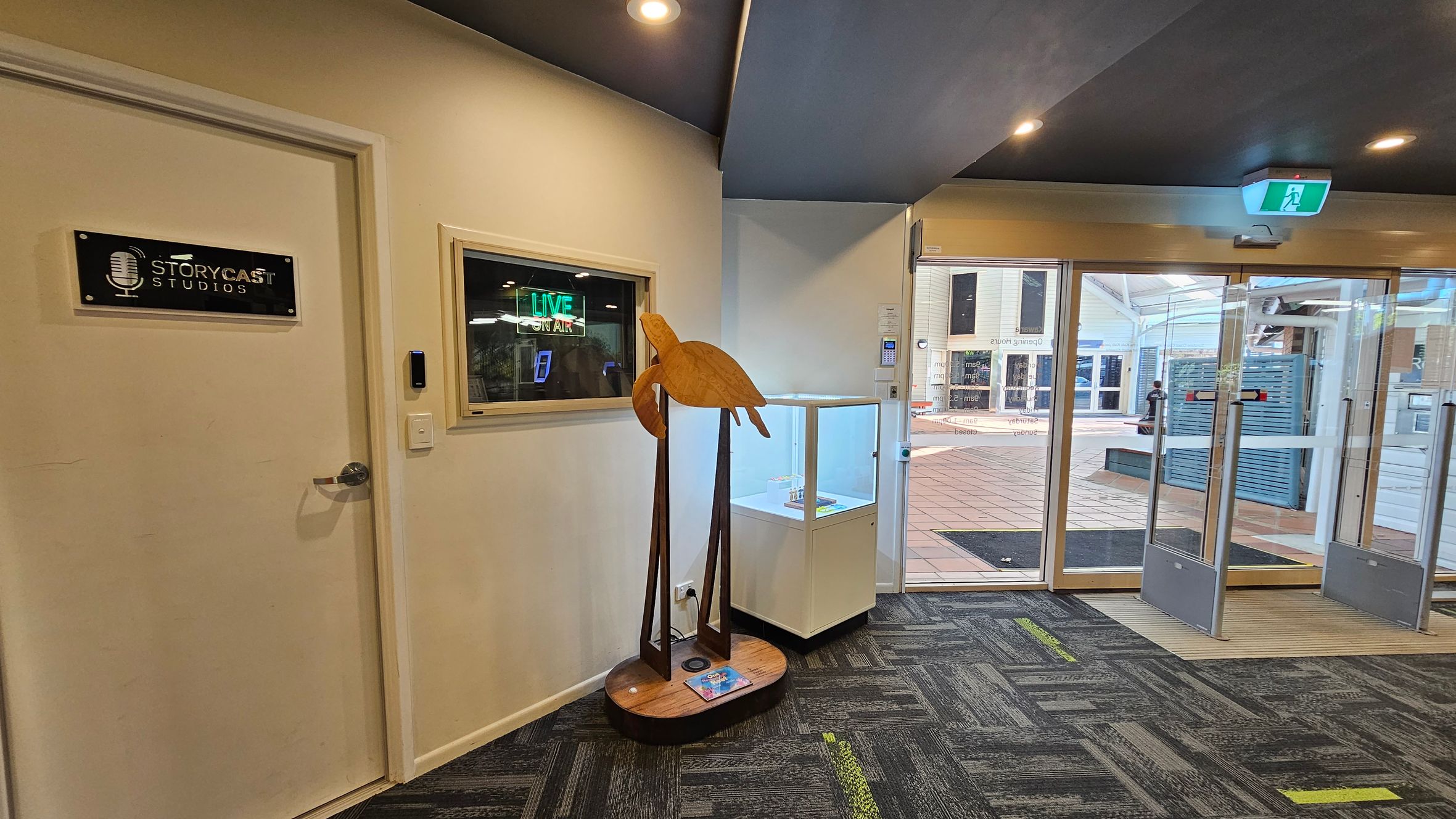 A hallway with a door on the left labelled "StoryCast Studios" and a window displaying a "LIVE ON AIR" sign. In the centre, there's a wooden sculpture of a bird on a stand. To the right, a glass display case is positioned near a set of glass doors leading outside. The floor is covered with a patterned carpet in shades of grey and green, and the ceiling is dark with recessed lighting. An illuminated exit sign is above the doors. A hallway with a door on the left labelled "StoryCast Studios" and a window displaying a "LIVE ON AIR" sign. In the centre, there's a wooden sculpture of a bird on a stand. To the right, a glass display case is positioned near a set of glass doors leading outside. The floor is covered with a patterned carpet in shades of grey and green, and the ceiling is dark with recessed lighting. An illuminated exit sign is above the doors.
