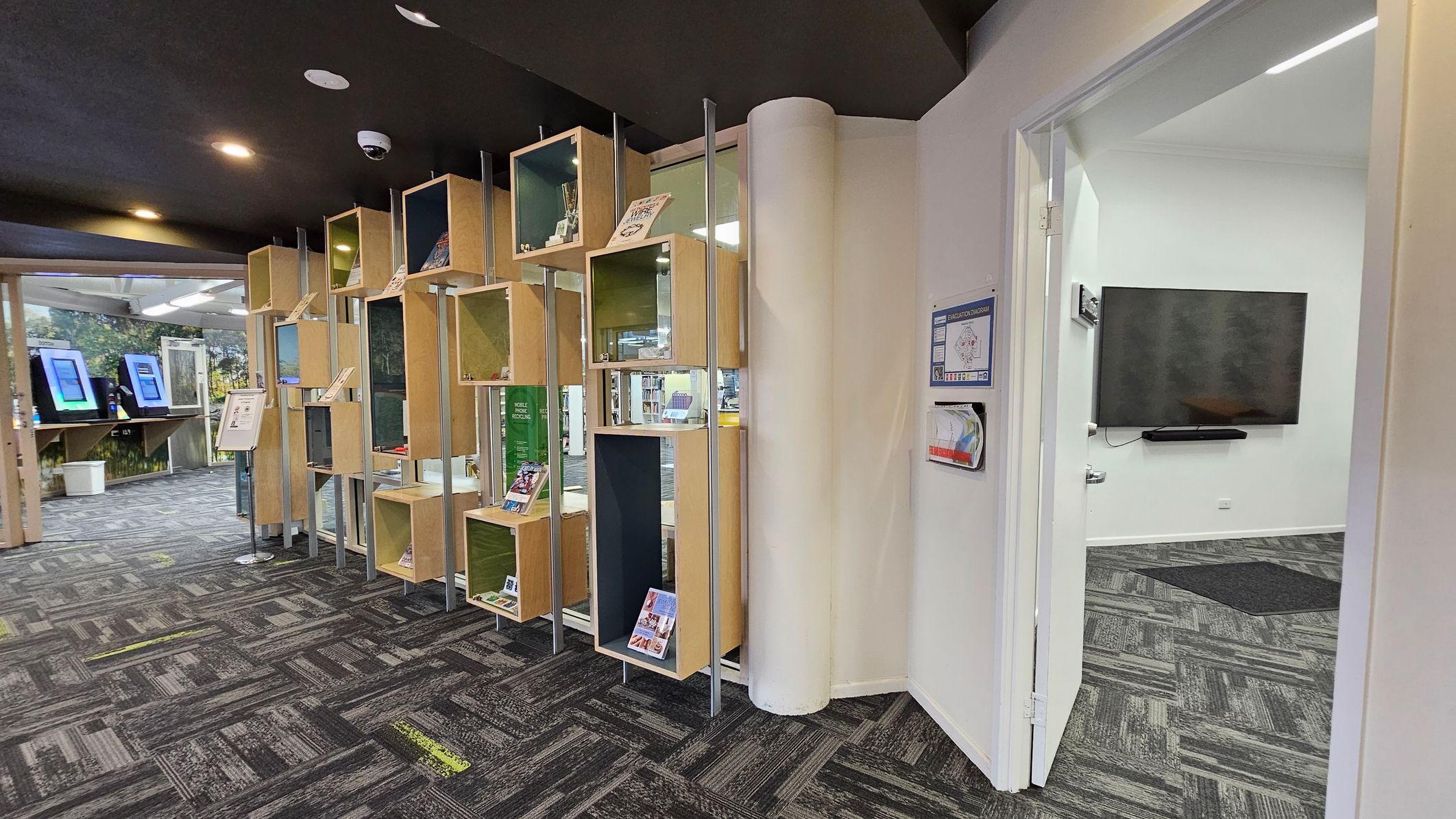 On the left, there are wooden display shelves with various items and books. Behind them, a section with digital screens is visible. The floor is carpeted with a grey and green pattern. On the right, an open door leads to a room with a large wall-mounted TV. The ceiling is dark with recessed lighting. On the left, there are wooden display shelves with various items and books. Behind them, a section with digital screens is visible. The floor is carpeted with a grey and green pattern. On the right, an open door leads to a room with a large wall-mounted TV. The ceiling is dark with recessed lighting.
